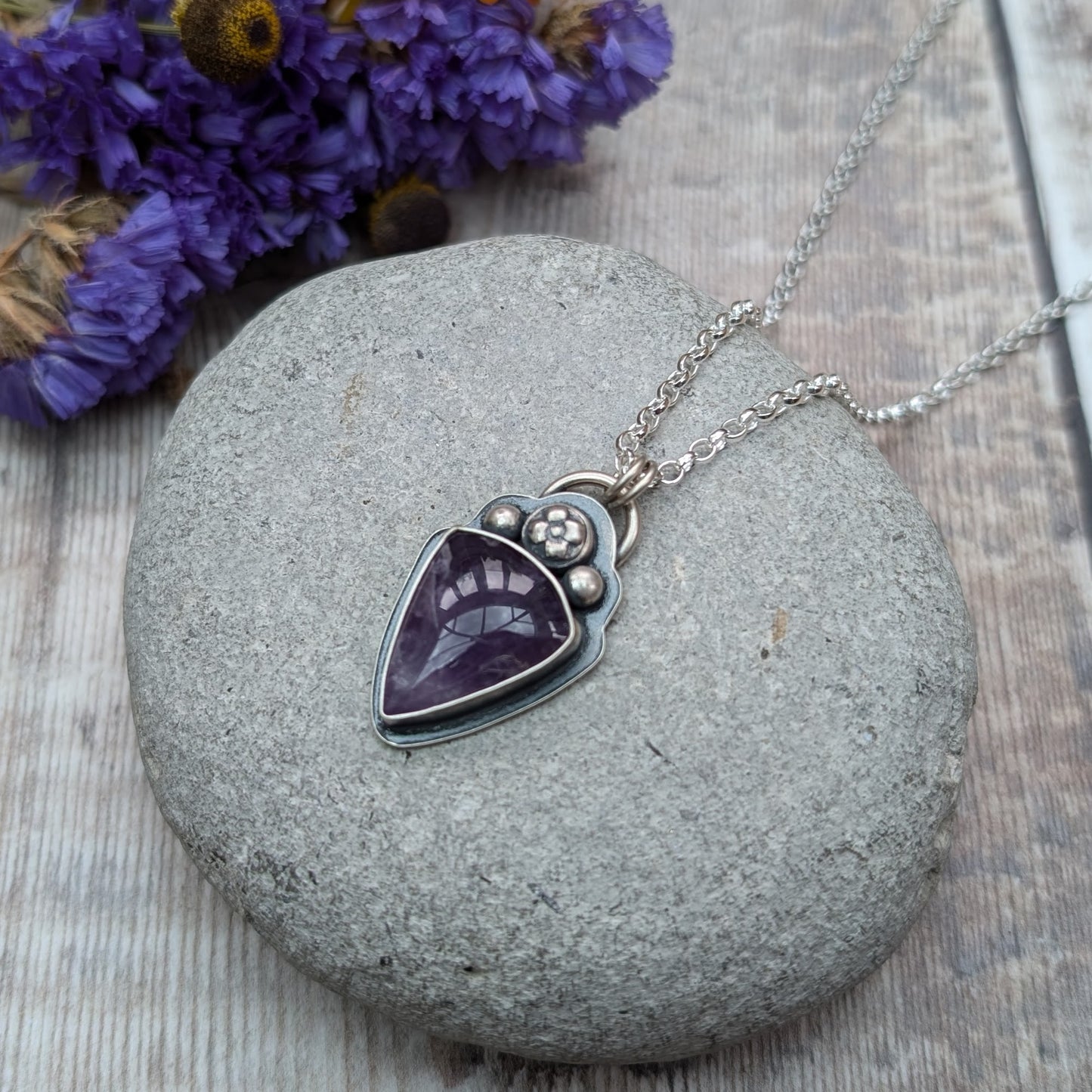 An oxidised silver necklace with a teardrop-shaped amethyst stone pendant. The pendant features a flower and bead design above the dark purple stone on the bezel setting.