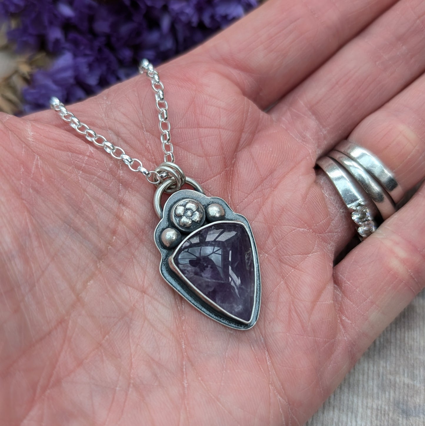 An oxidised silver necklace with a teardrop-shaped amethyst stone pendant. The pendant features a flower and bead design above the dark purple stone on the bezel setting.