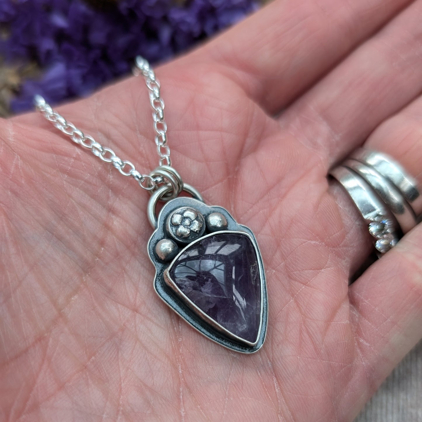 An oxidised silver necklace with a teardrop-shaped amethyst stone pendant. The pendant features a flower and bead design above the dark purple stone on the bezel setting.