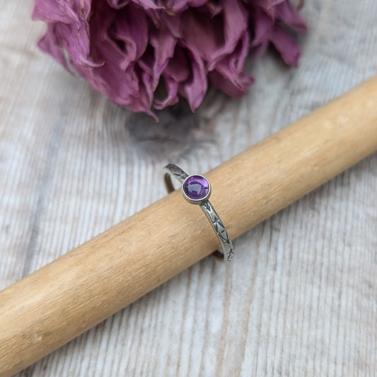 Slim silver band with cross stamped pattern. A purple Amethyst round stone is mounted on the band in an oxidised sterling silver bezel. Rings can be stacked above or below this piece.