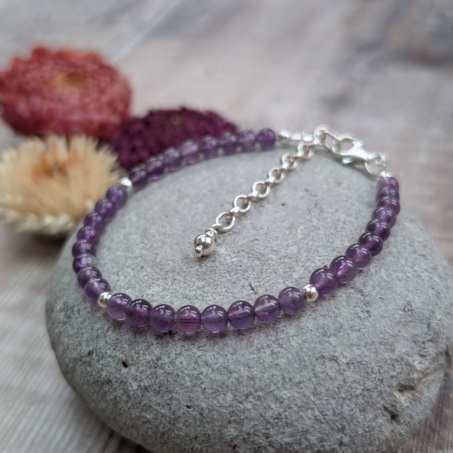 Amethyst bead bracelet with sterling silver spacer beads and extender chain, displayed on a smooth grey stone with dried flowers in the background.