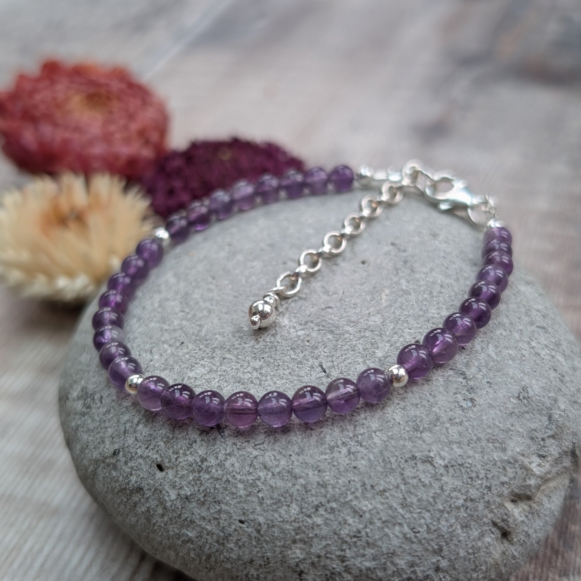 Amethyst bead bracelet with sterling silver spacer beads and extender chain, displayed on a smooth grey stone with dried flowers in the background.
