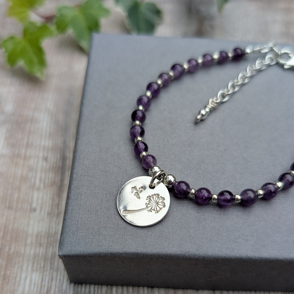 Close up of amethyst and silver dandelion disc bracelet on a grey jewellery box, highlighting the hand stamped dandelion design, purple gemstone beads and silver details.