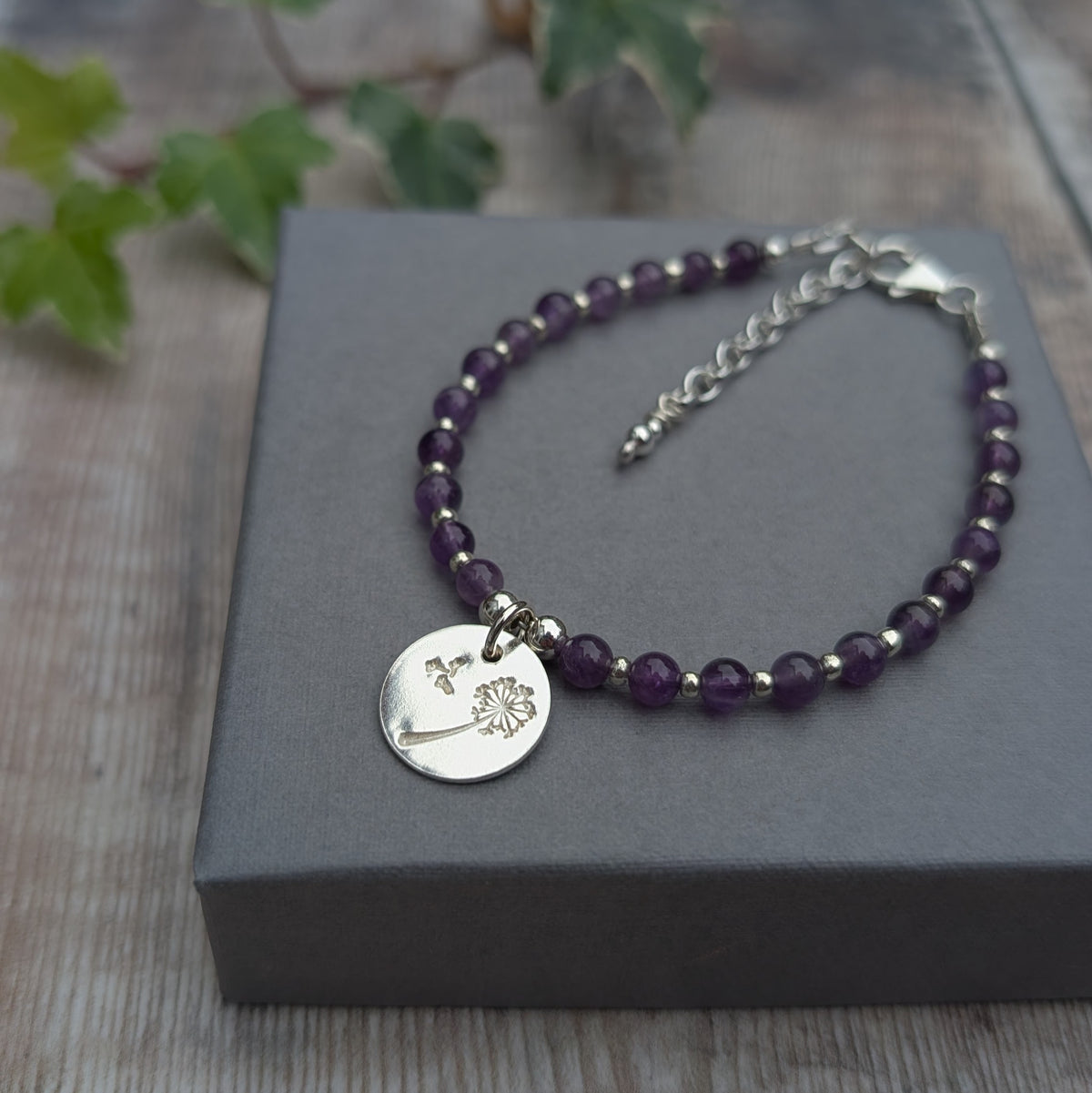 Amethyst and silver dandelion disc bracelet displayed on a grey jewellery box, showing purple amethyst beads, silver spacers and a hand stamped dandelion charm.