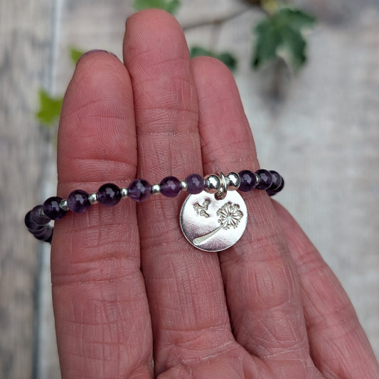 Close up of amethyst and silver dandelion disc bracelet held across fingers, showing the purple gemstone beads and shiny silver dandelion charm.