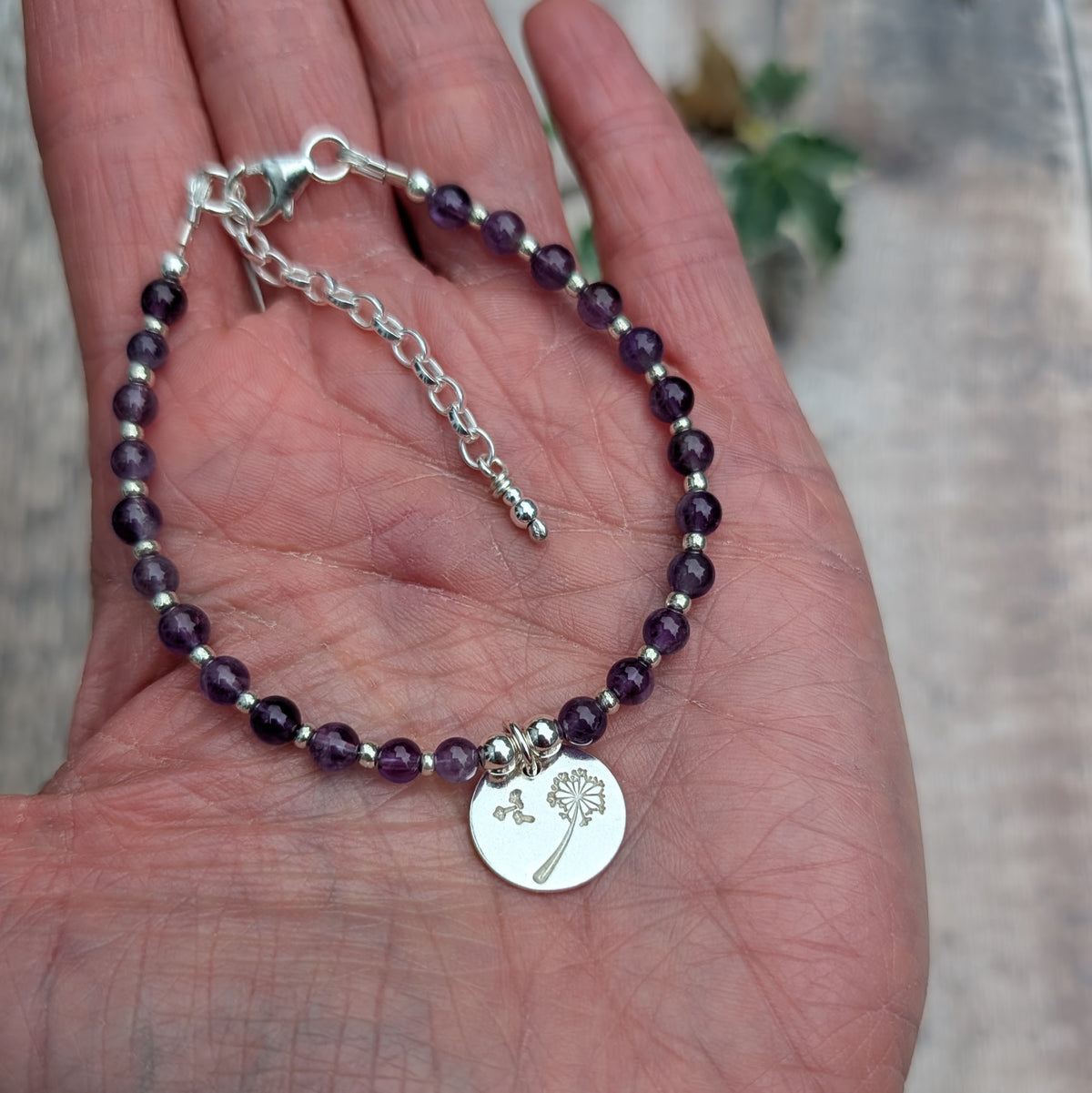 Amethyst and silver dandelion disc bracelet resting in the palm of a hand, showing the full bracelet, sterling silver clasp, extension chain and stamped dandelion charm.