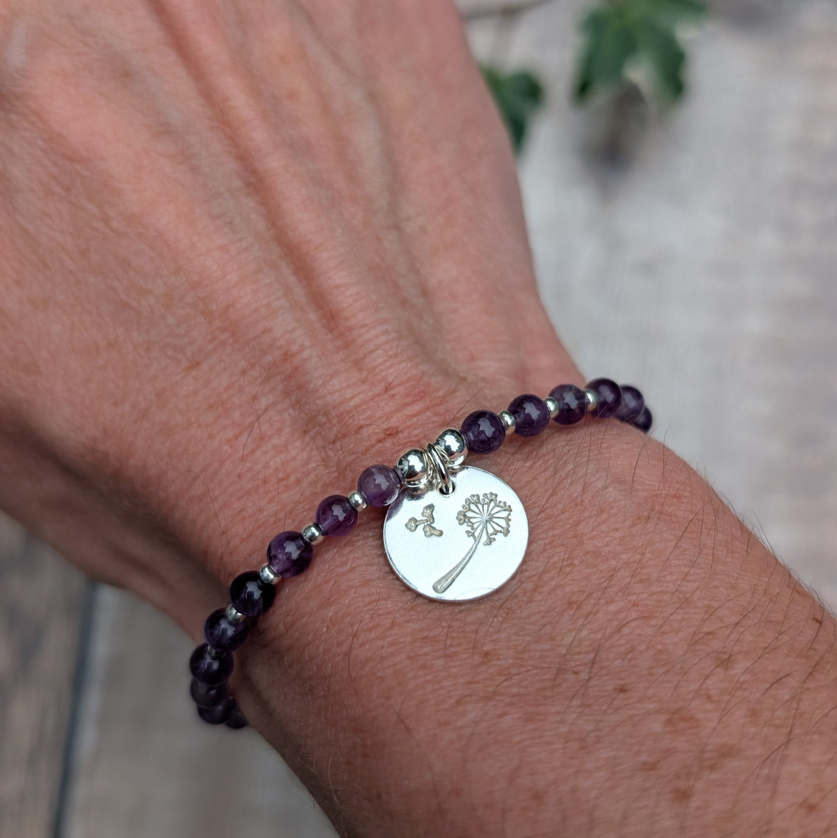 Amethyst and silver dandelion disc bracelet being worn on a wrist, showing the purple amethyst beads and round sterling silver dandelion charm.