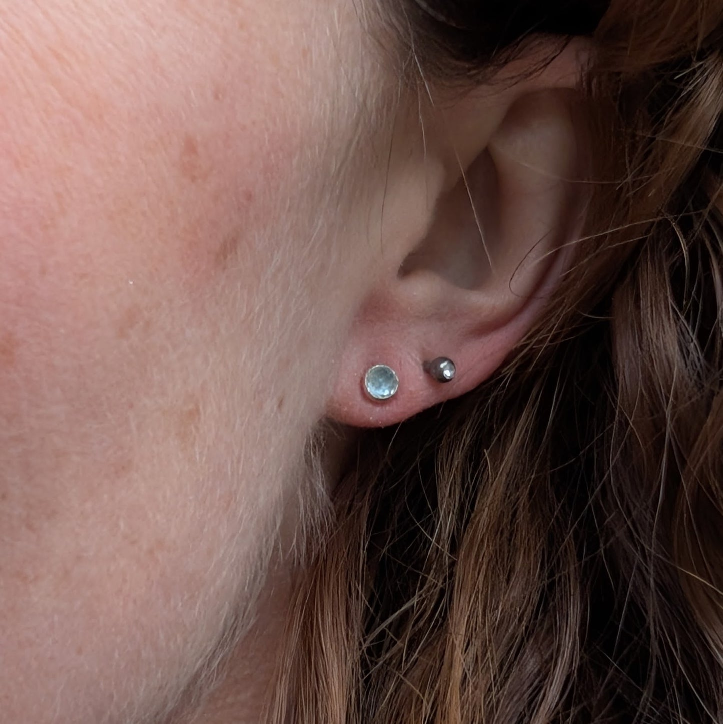 Each earring is a round rose cut Aquamarine gemstone set in silver bezel attached to stud bar. Stone is pale blue translucent with uneven rose cut finish that catches the light at different angles.