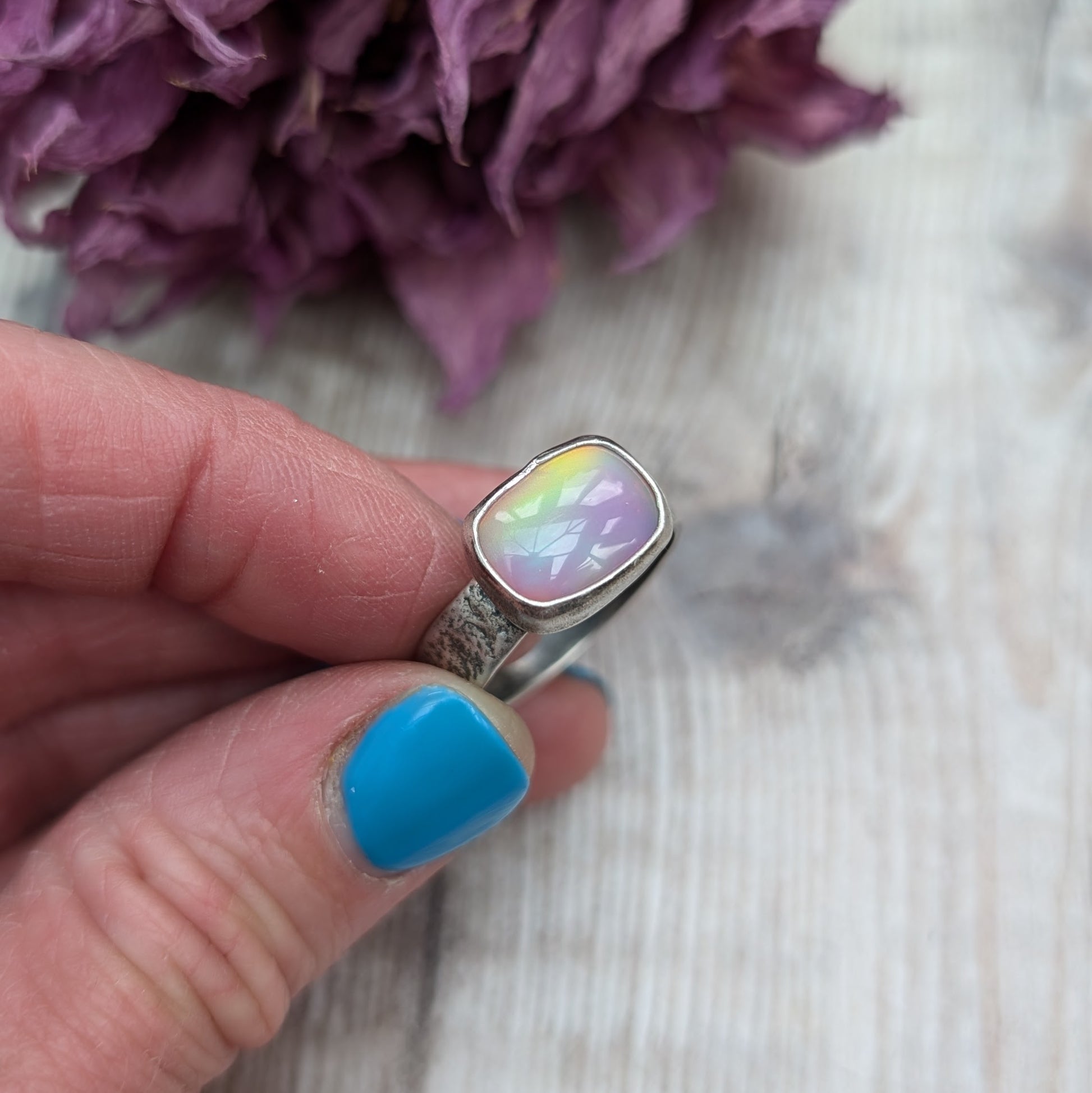 An oxidised silver ring with textured band approx. 5mm wide featuring an iridescent, rectangular opal stone. The opal reflects a range of colors, predominantly pink and yellow.