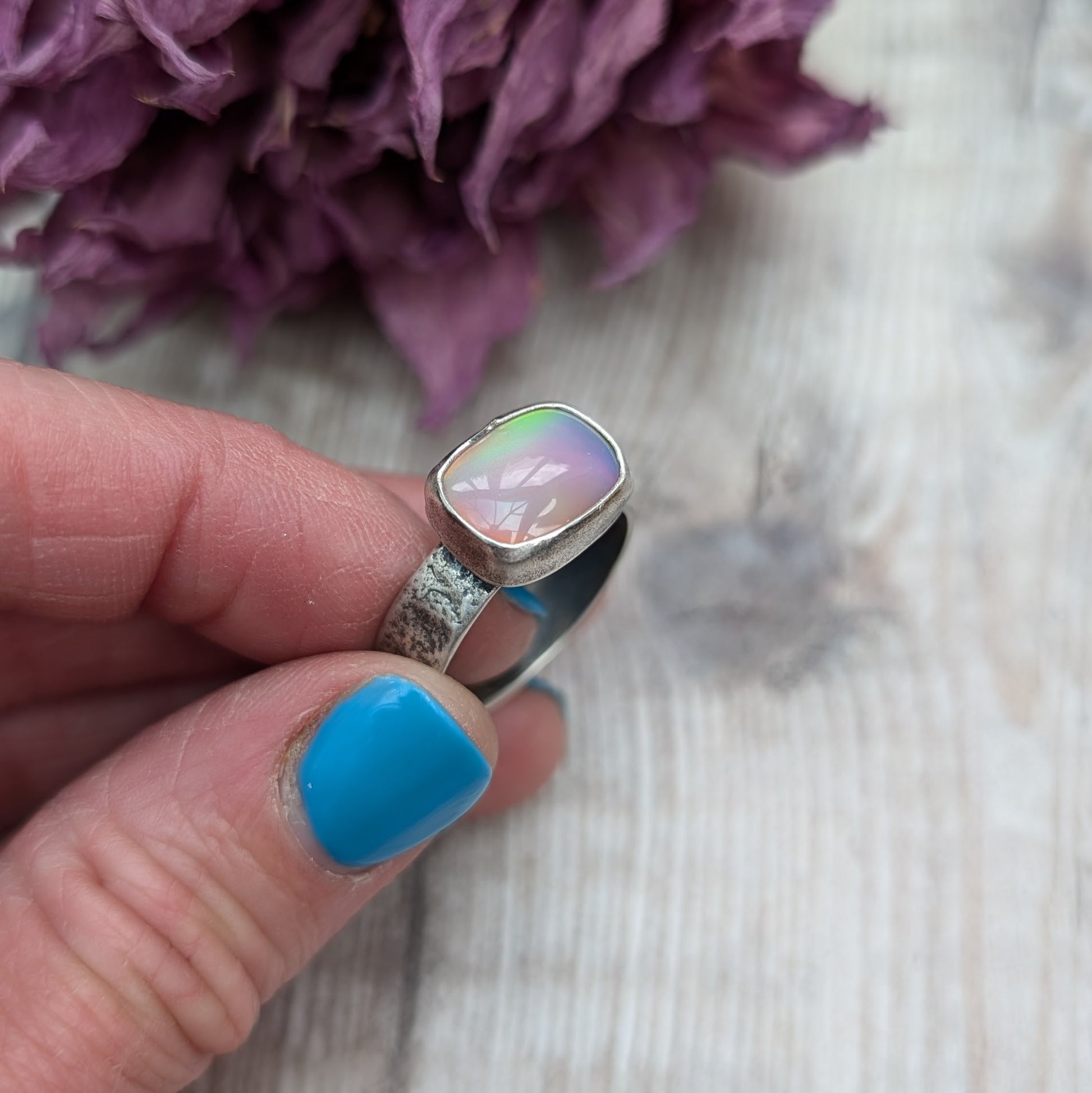 An oxidised silver ring with textured band approx. 5mm wide featuring an iridescent, rectangular opal stone. The opal reflects a range of colors, predominantly pink and yellow.