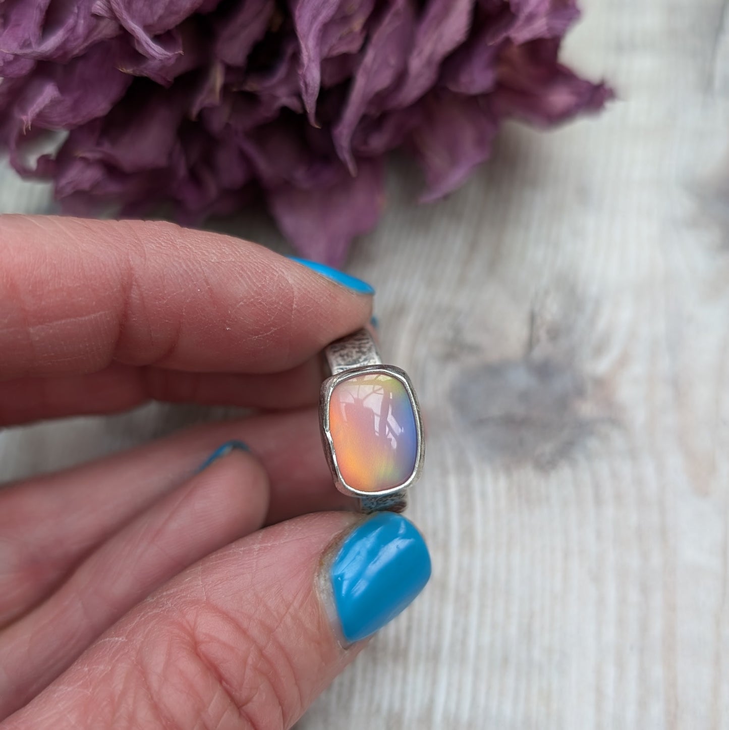 An oxidised silver ring with textured band approx. 5mm wide featuring an iridescent, rectangular opal stone. The opal reflects a range of colors, predominantly pink and yellow.