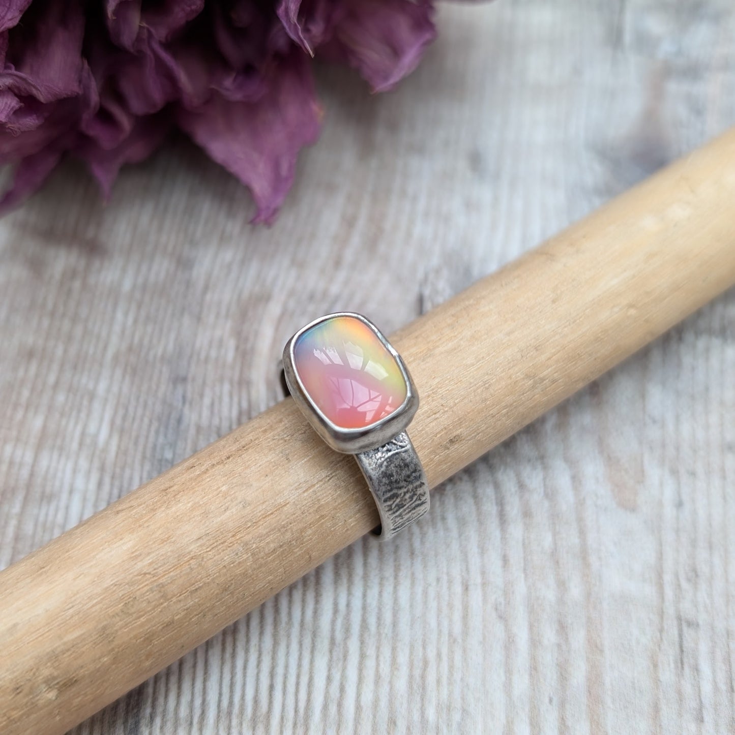 An oxidised silver ring with textured band approx. 5mm wide featuring an iridescent, rectangular opal stone. The opal reflects a range of colors, predominantly pink and yellow.