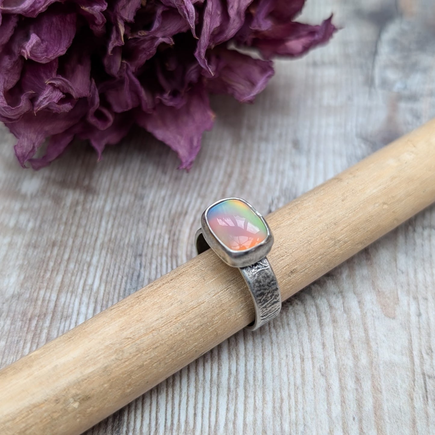 An oxidised silver ring with textured band approx. 5mm wide featuring an iridescent, rectangular opal stone. The opal reflects a range of colors, predominantly pink and yellow.