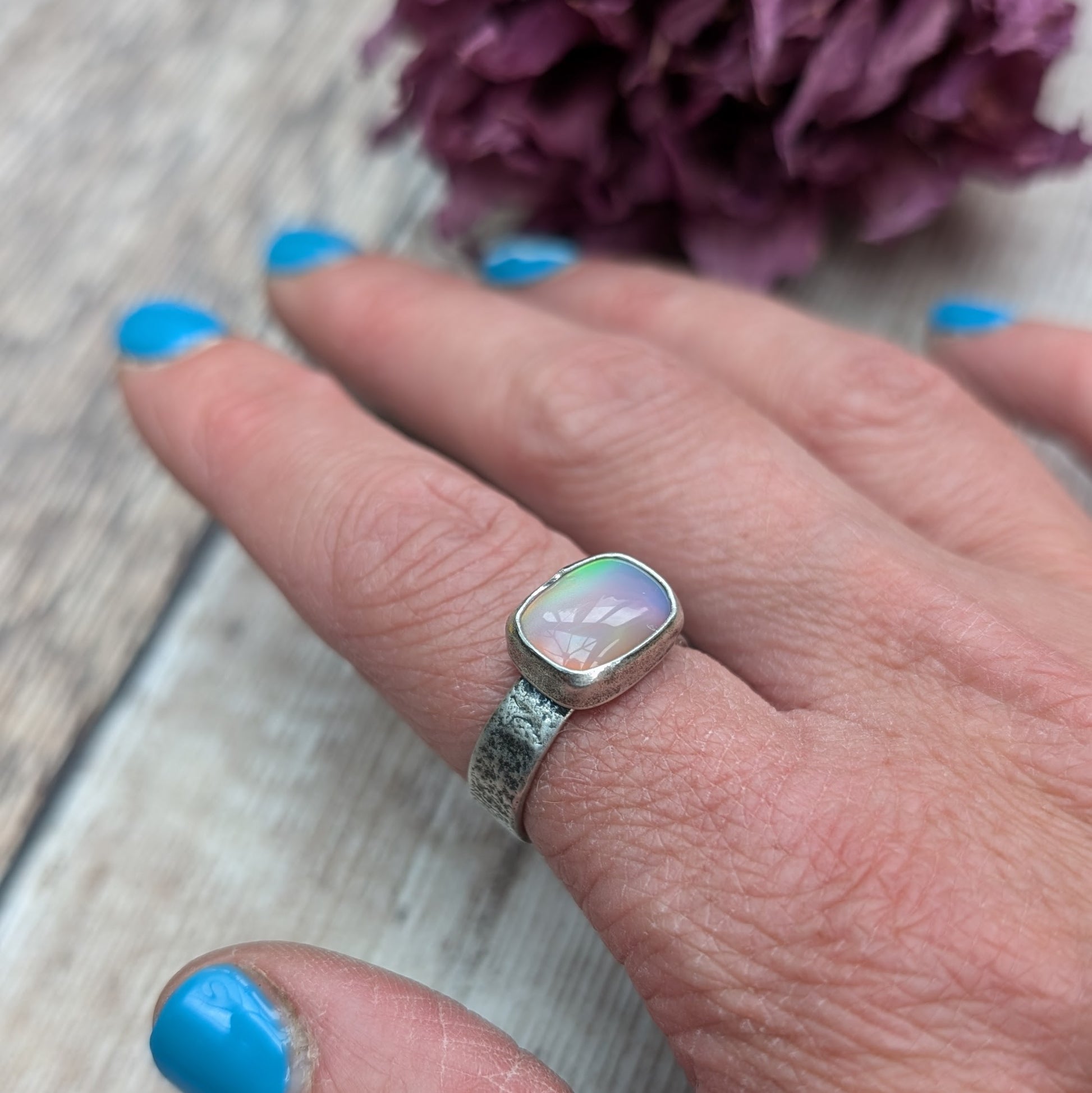 An oxidised silver ring with textured band approx. 5mm wide featuring an iridescent, rectangular opal stone. The opal reflects a range of colors, predominantly pink and yellow.