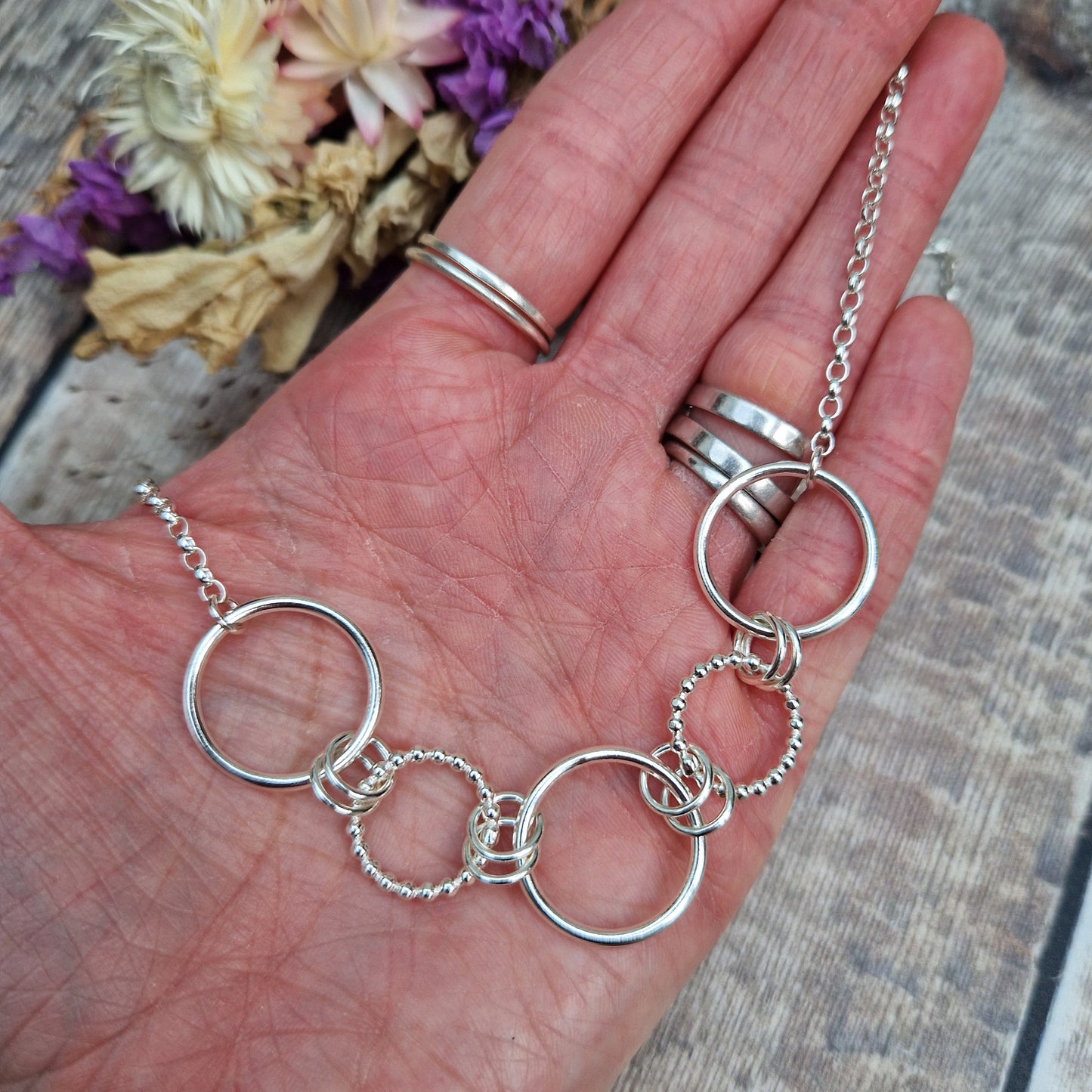 Five open silver circles in a pattern of smooth finish linked to beaded circle linked to smooth finish linked to beaded circle linked to smooth finish. Each linked in a line with each other via two small silver circles. Each smooth circle at each end of the pattern is attached to a silver chain.