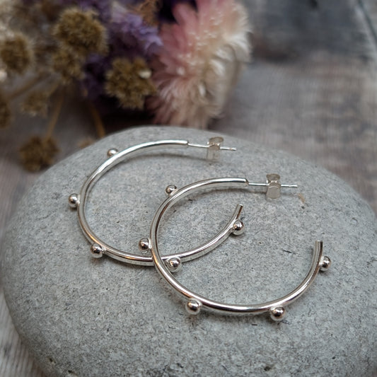 Pair of silver hoop earrings with small pebble-like details evenly spaced around the hoop, placed on a smooth stone with dried flowers in the background