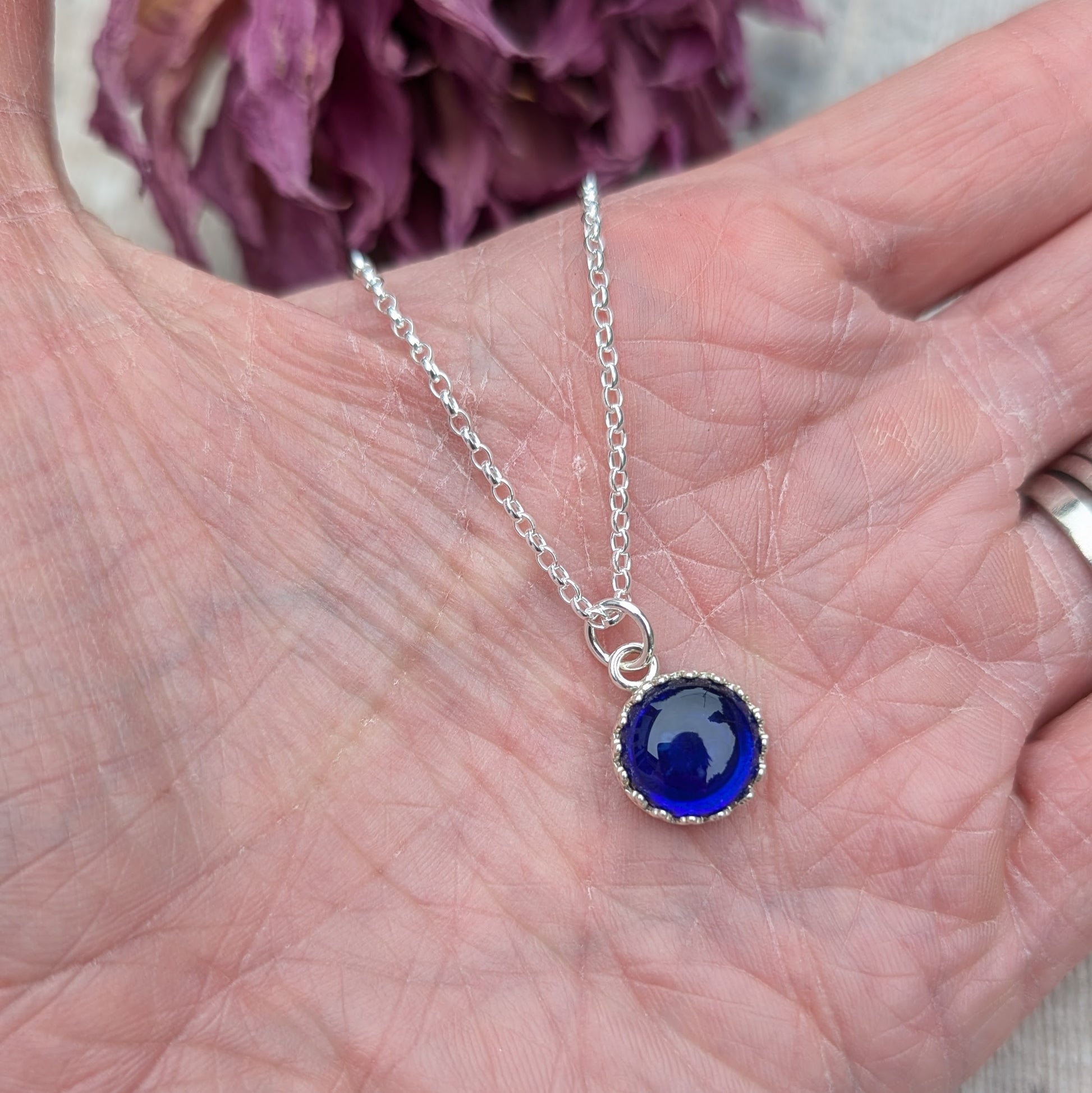 Pendant is a deep blue petite glass stone in circle shape, set in an intricate pattered Sterling Silver bezel, suspended from a silver chain via a small open circle link.