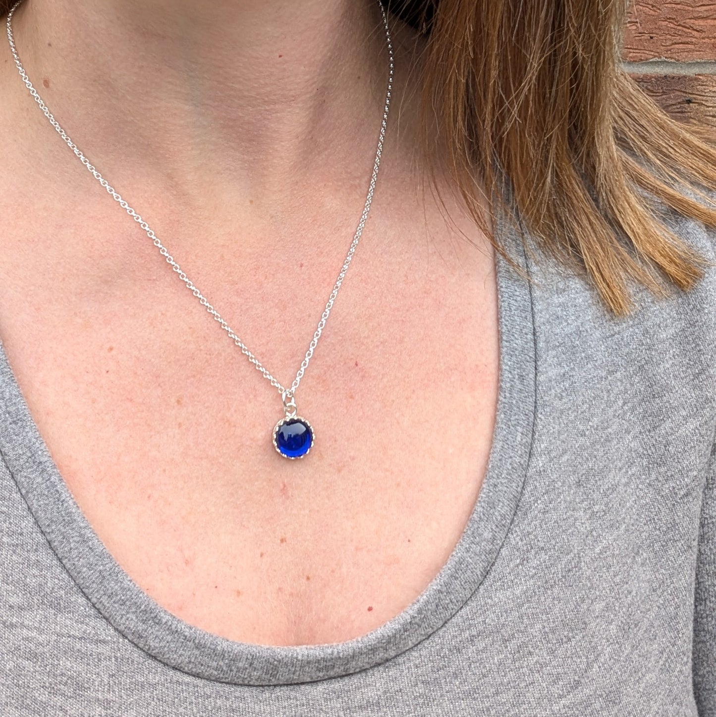 Pendant is a deep blue petite glass stone in circle shape, set in an intricate pattered Sterling Silver bezel, suspended from a silver chain via a small open circle link.