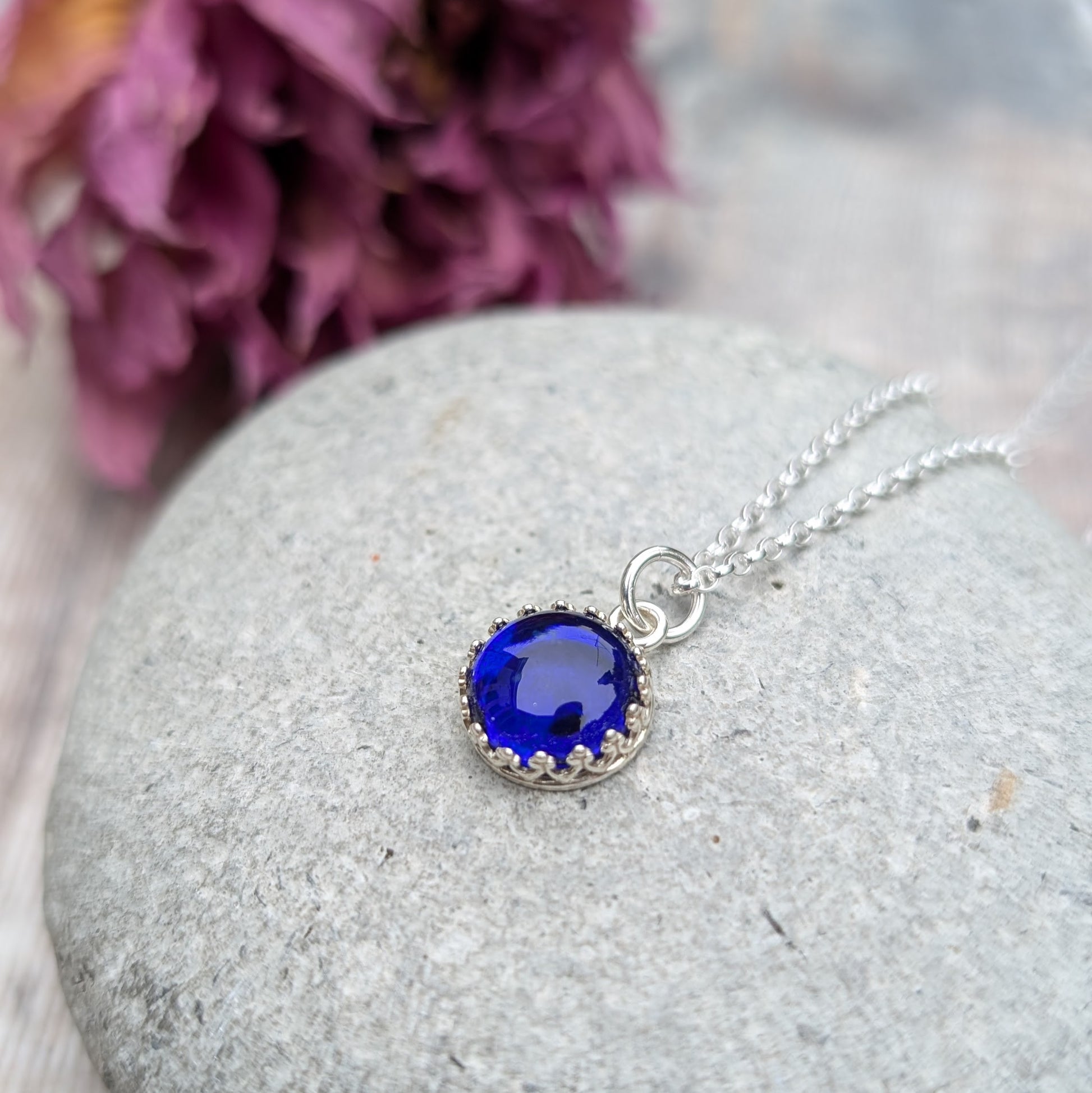 Pendant is a deep blue petite glass stone in circle shape, set in an intricate pattered Sterling Silver bezel, suspended from a silver chain via a small open circle link.