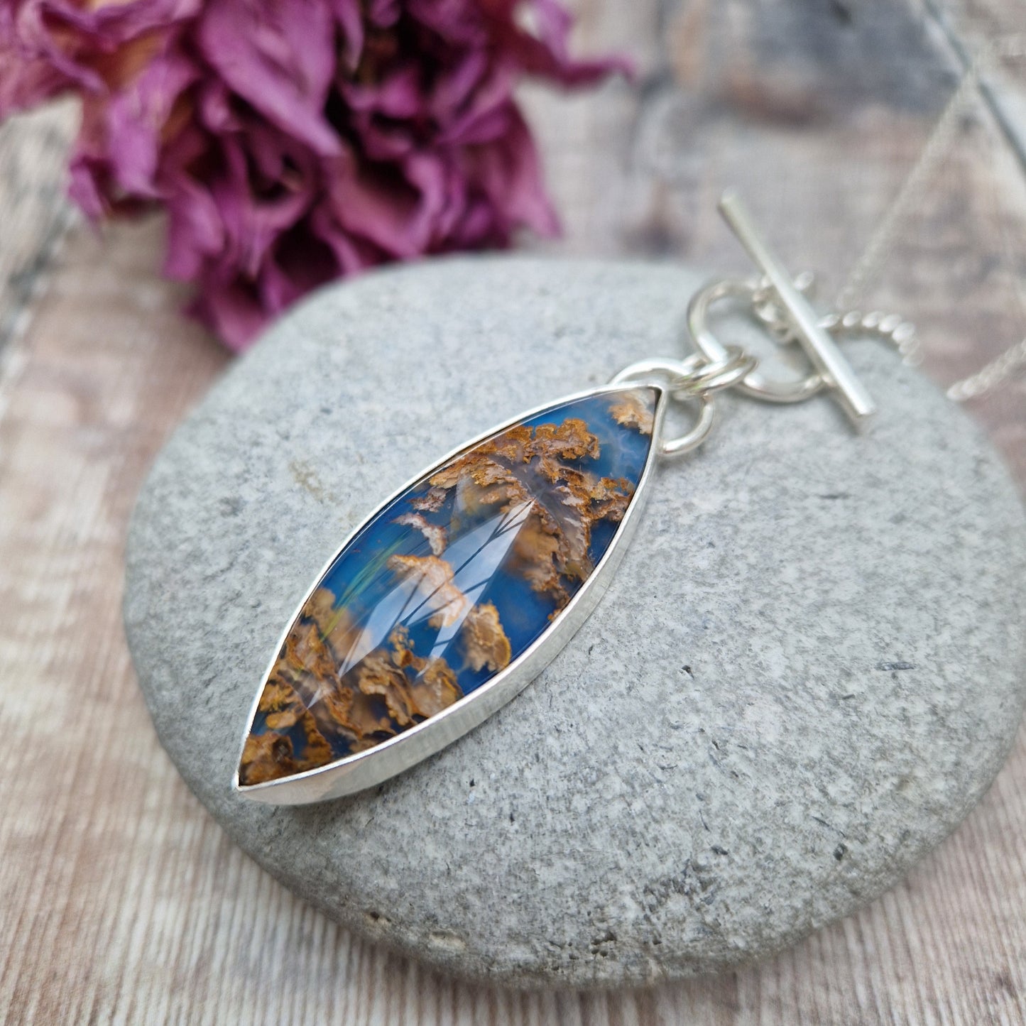 Marquise shaped pendant 45mm long set in silver bezel. Agate stone is cornflower blue with flecks of brown and gold plumes. Pendant is attached to a Sterling Silver chain via two small silver circle links and a t-bar through larger circle link.