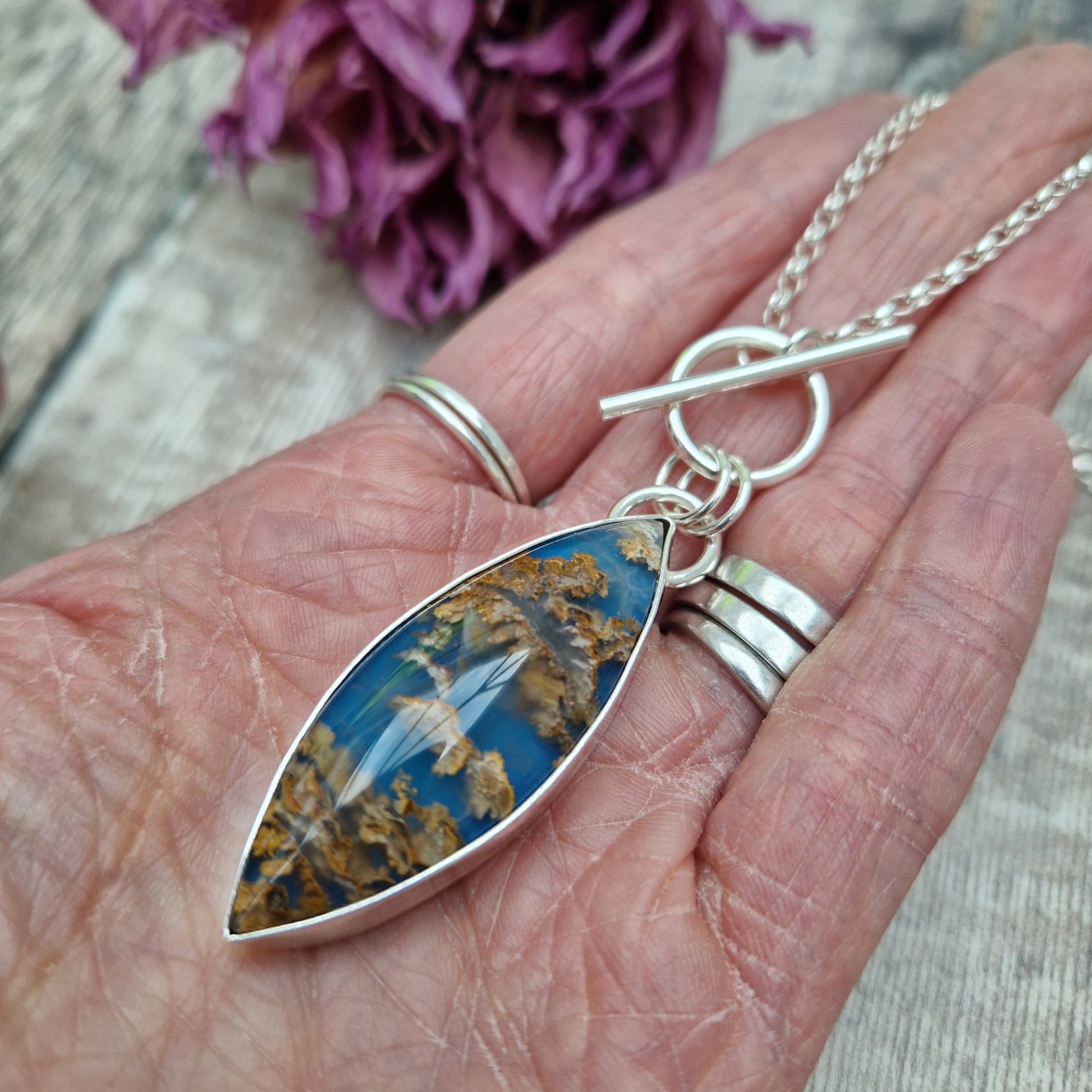 Marquise shaped pendant 45mm long set in silver bezel. Agate stone is cornflower blue with flecks of brown and gold plumes. Pendant is attached to a Sterling Silver chain via two small silver circle links and a t-bar through larger circle link.