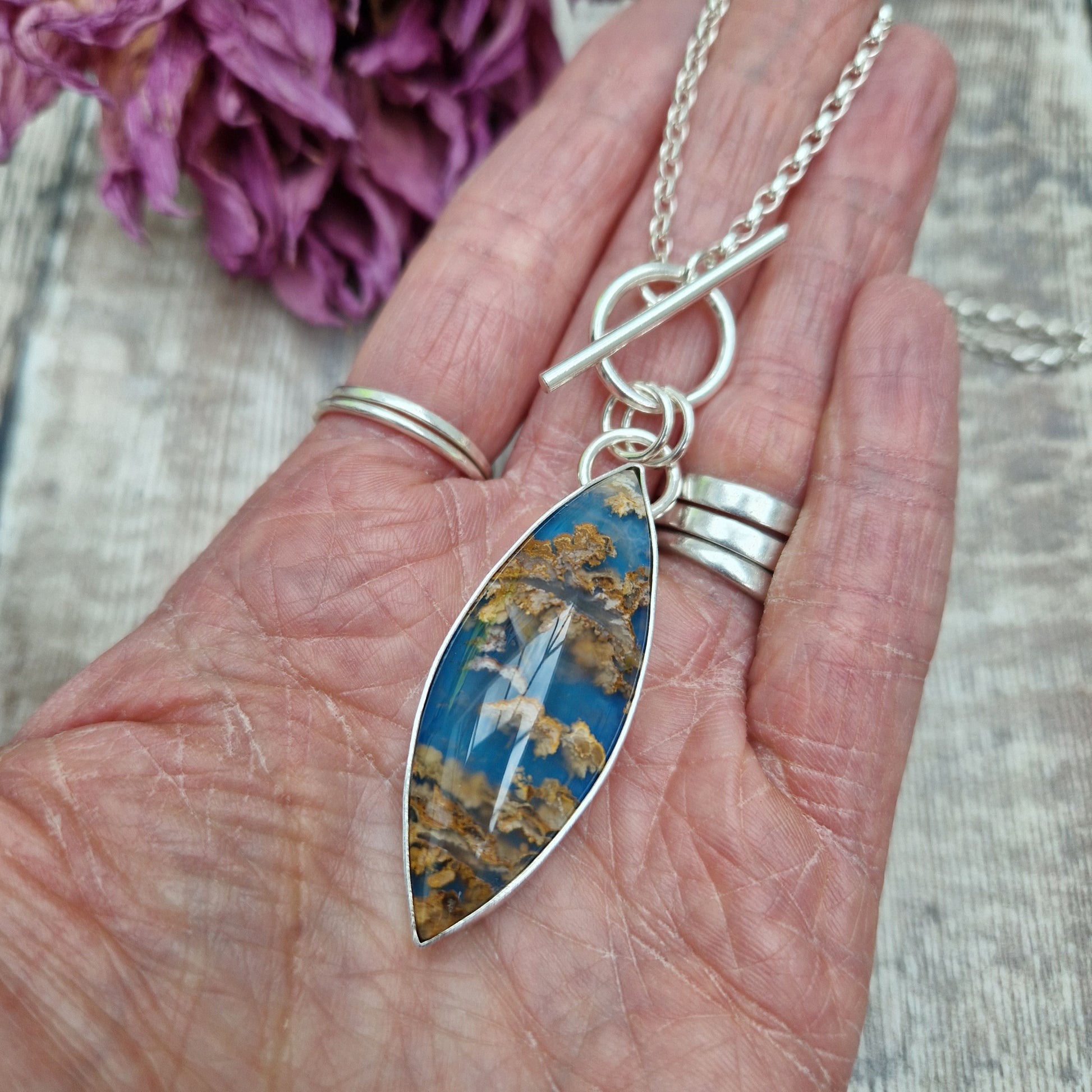 Marquise shaped pendant 45mm long set in silver bezel. Agate stone is cornflower blue with flecks of brown and gold plumes. Pendant is attached to a Sterling Silver chain via two small silver circle links and a t-bar through larger circle link.