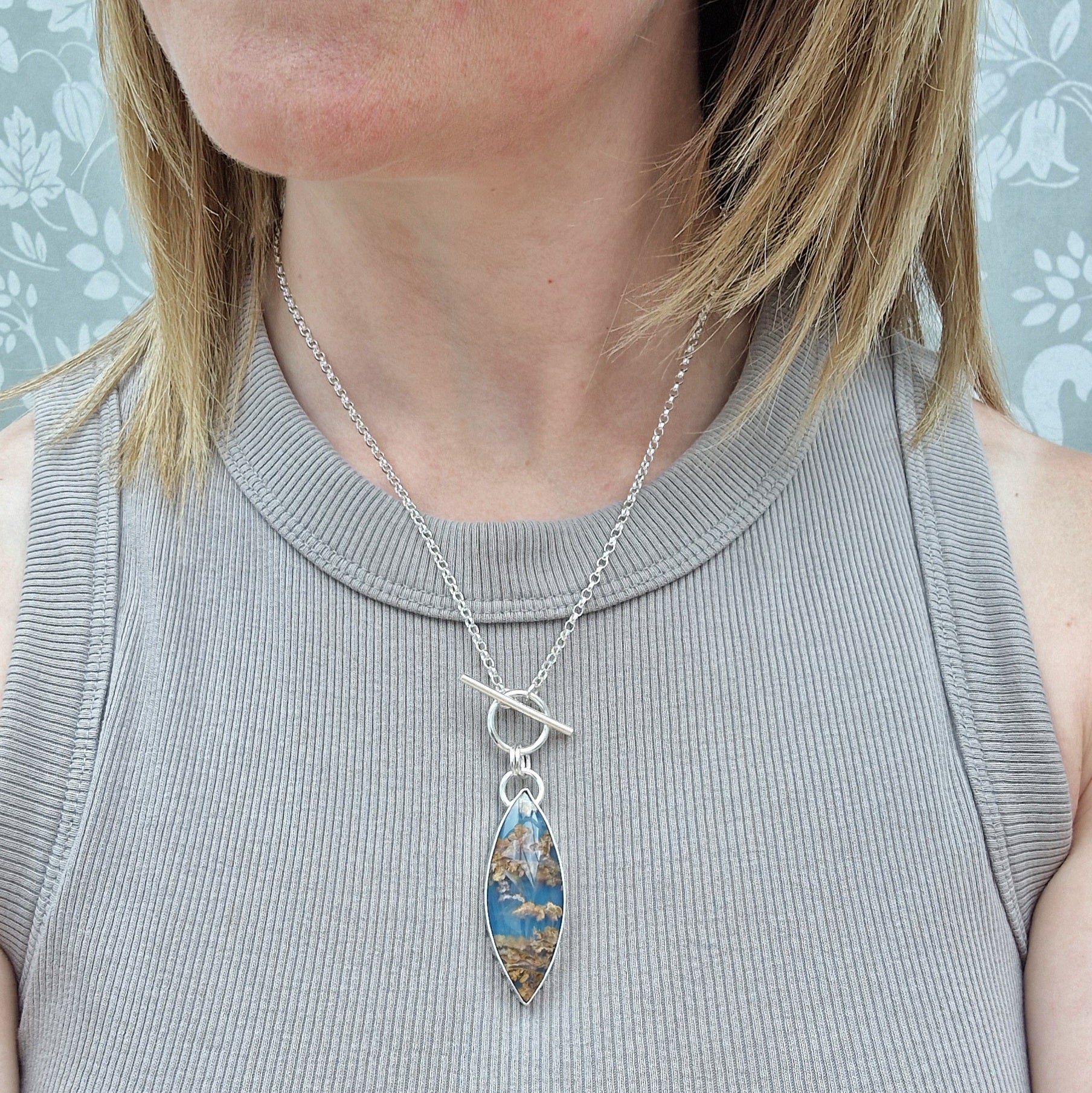 Marquise shaped pendant 45mm long set in silver bezel. Agate stone is cornflower blue with flecks of brown and gold plumes. Pendant is attached to a Sterling Silver chain via two small silver circle links and a t-bar through larger circle link.