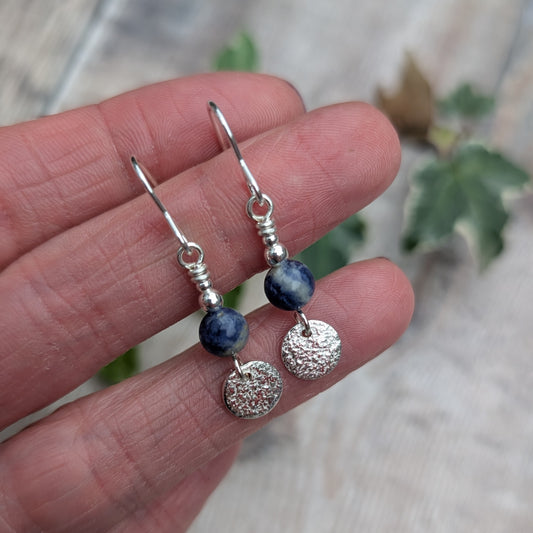 Close up of blue sodalite and silver textured disc earrings resting on fingers, showing the natural blue and white patterns of the sodalite beads, textured sterling silver discs and silver bead details.