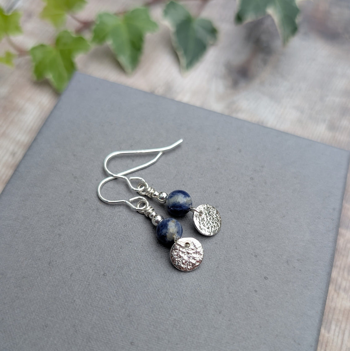 Blue sodalite and silver textured disc earrings handmade from recycled sterling silver, featuring round blue gemstone beads and small textured silver discs, displayed on a grey background.