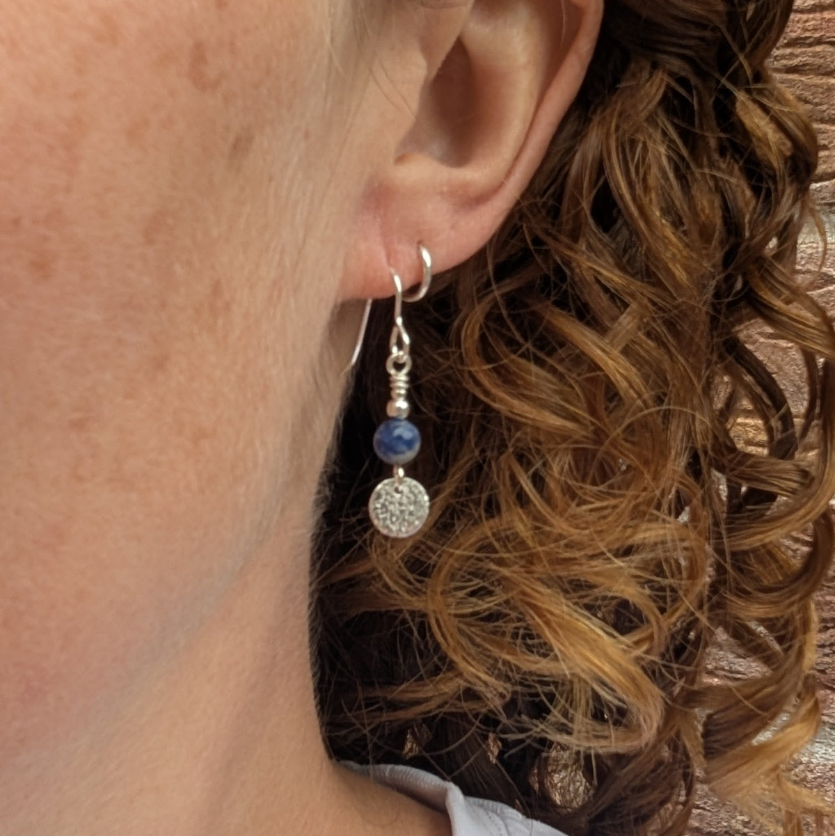 Blue sodalite and silver textured disc earrings being worn, showing the deep blue sodalite beads, handmade silver ear wires and small textured silver discs hanging below.