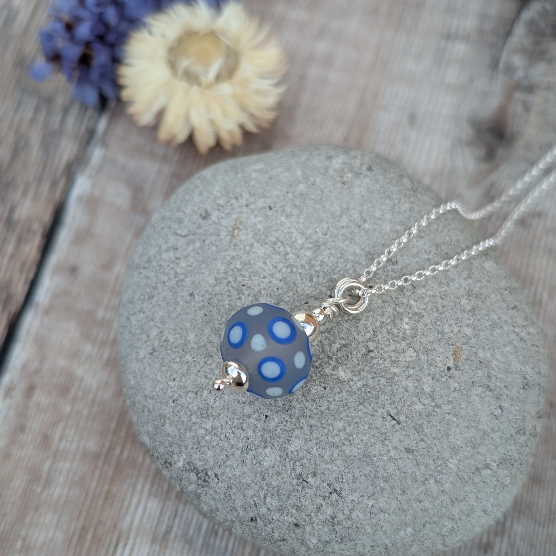 Blue Spotty Lampwork Glass Necklace resting on a smooth stone with dried flowers in the background.