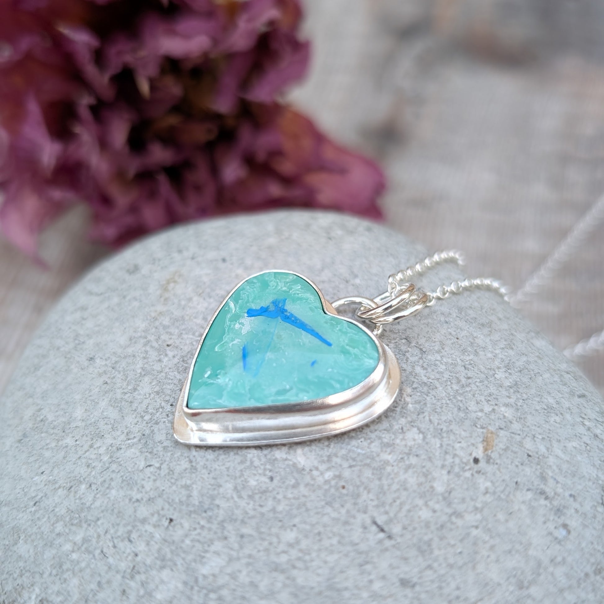 Pendant is blue heart shaped surfite set in a silver bezel surround, suspended from chain via two small open circle rings. Reverse of pendant has slightly raised heart shape stamped with floral pattern. Surfite is pale blue turquoise with white and darker blue flecks.