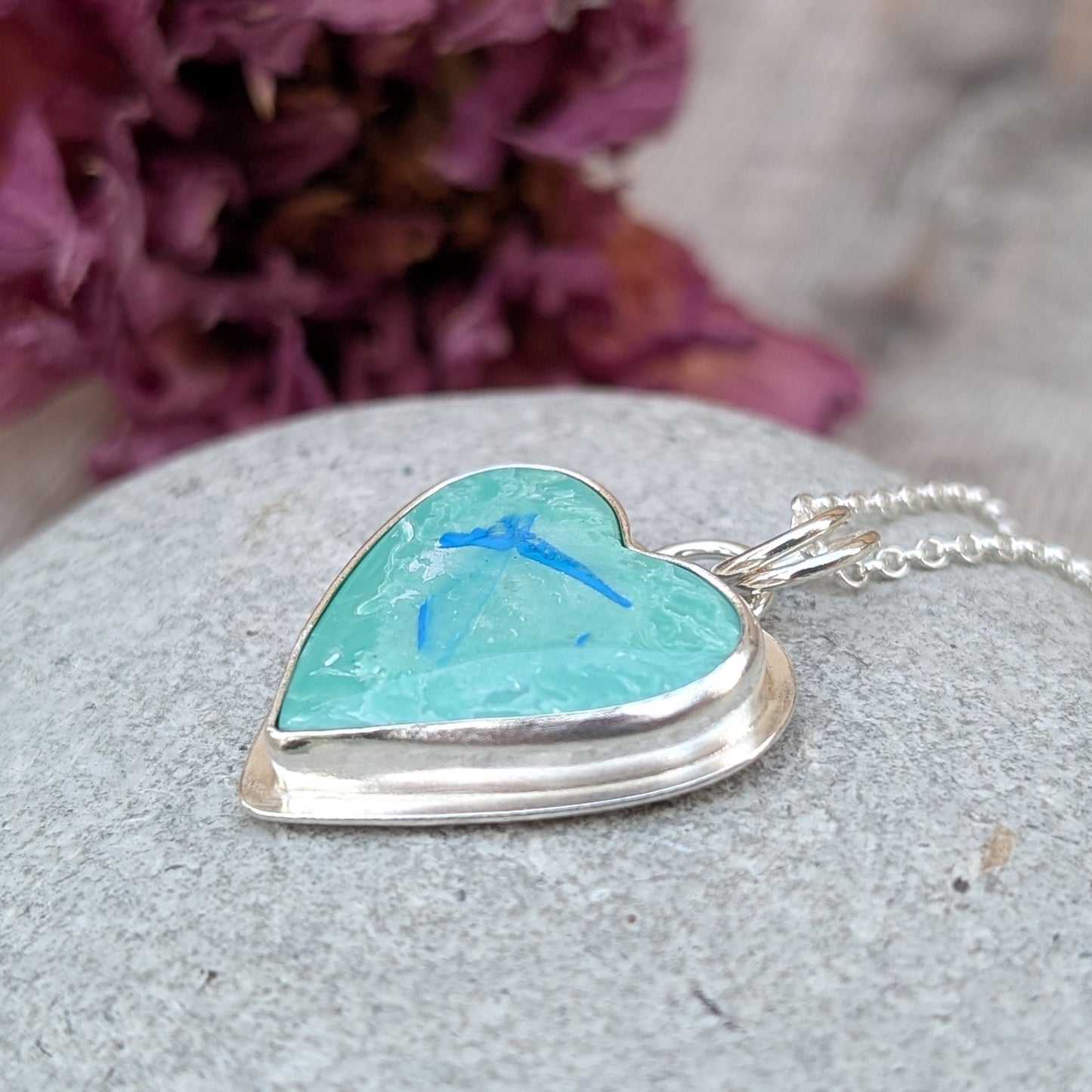 Pendant is blue heart shaped surfite set in a silver bezel surround, suspended from chain via two small open circle rings. Reverse of pendant has slightly raised heart shape stamped with floral pattern. Surfite is pale blue turquoise with white and darker blue flecks.