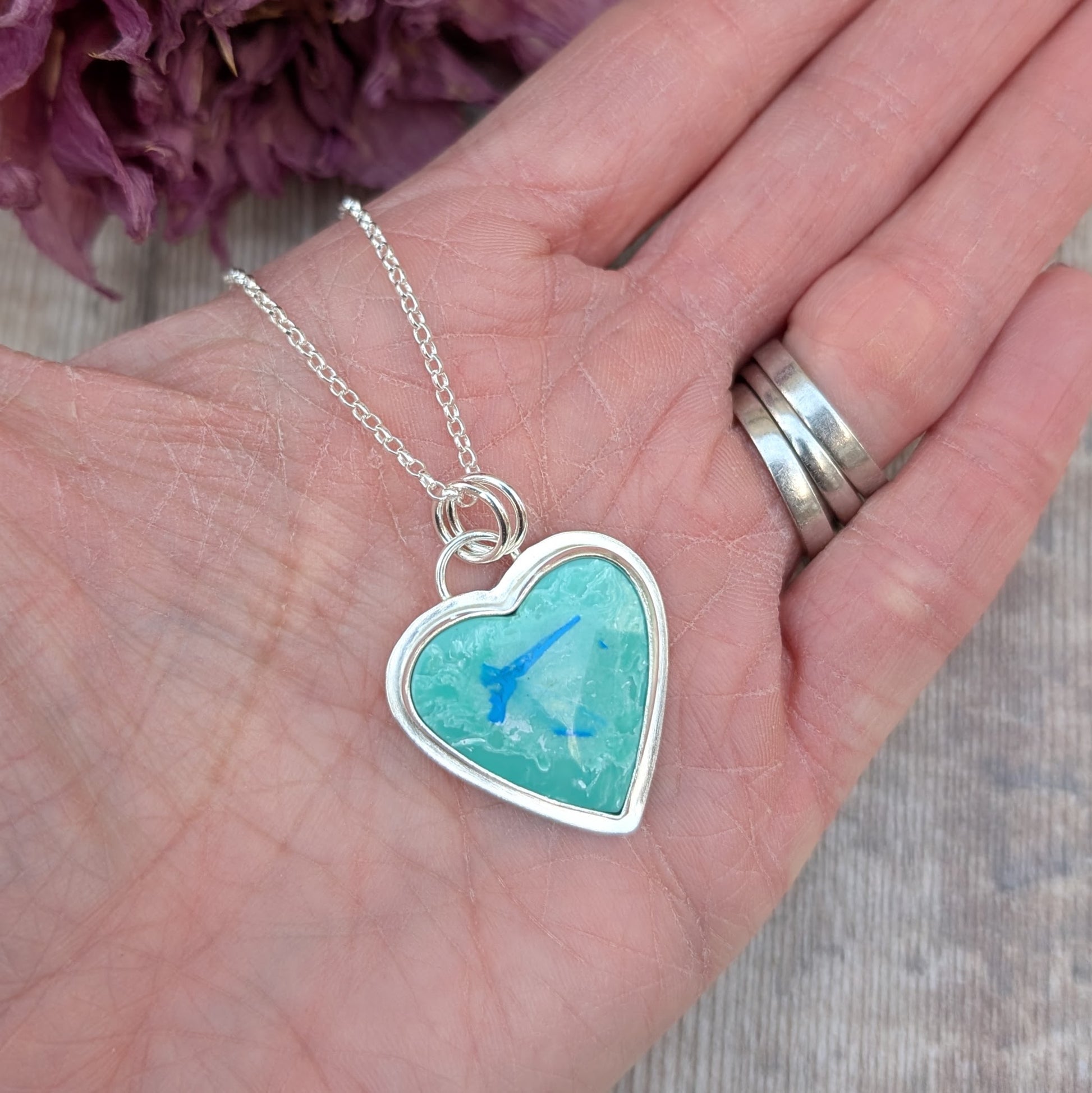 Pendant is blue heart shaped surfite set in a silver bezel surround, suspended from chain via two small open circle rings. Reverse of pendant has slightly raised heart shape stamped with floral pattern. Surfite is pale blue turquoise with white and darker blue flecks.