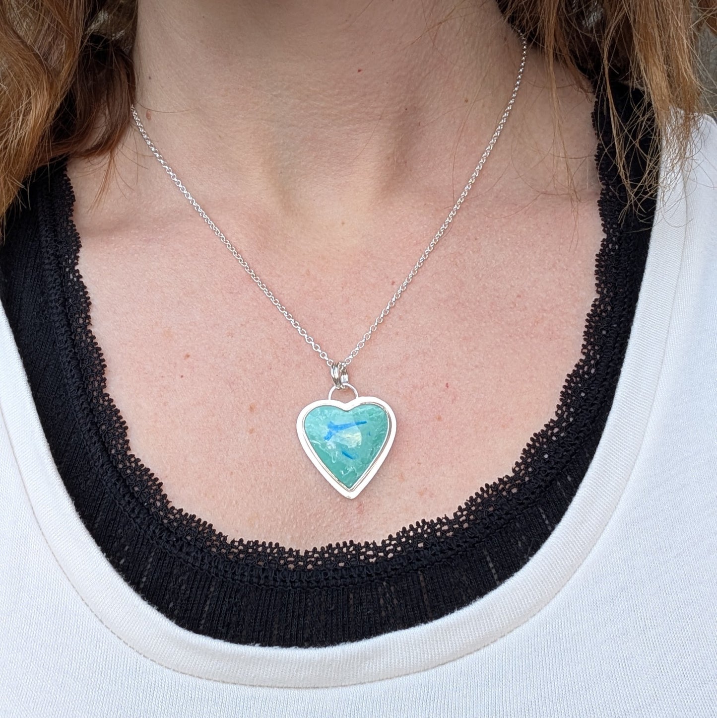 Pendant is blue heart shaped surfite set in a silver bezel surround, suspended from chain via two small open circle rings. Reverse of pendant has slightly raised heart shape stamped with floral pattern. Surfite is pale blue turquoise with white and darker blue flecks.