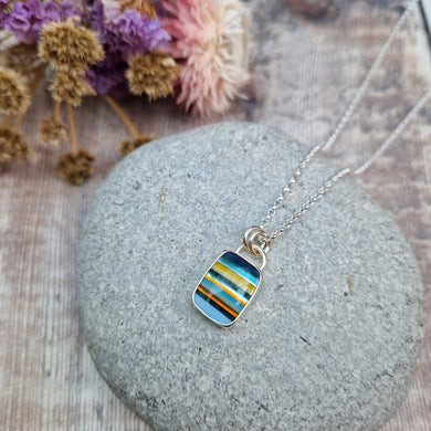 Rectangular shaped surfite pendant set in silver bezel. The stone has horizonal stripes in blues, yellows, orange and dark brown, slightly raised with glossy finish. Attached to silver chain via two small circular silver links.