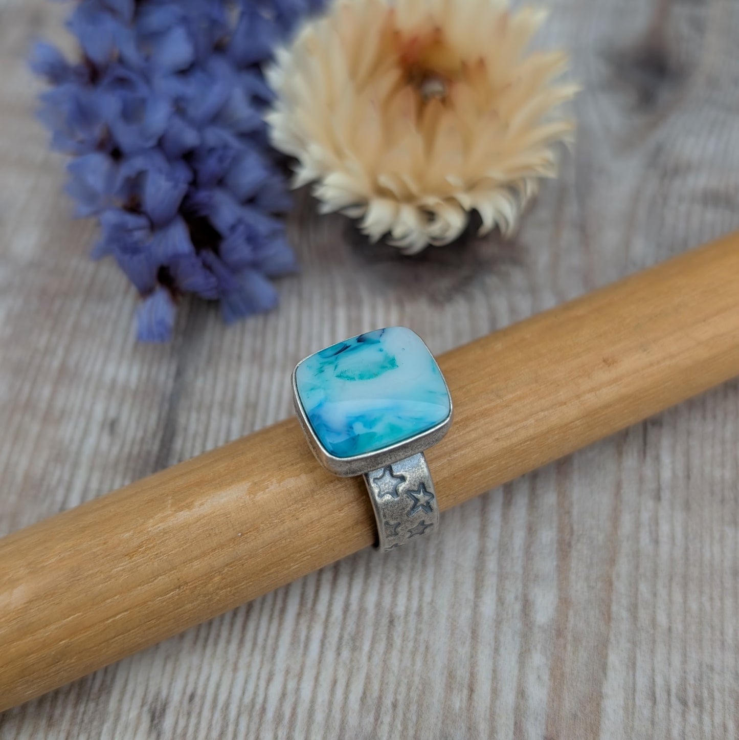 Blue Surfite Rectangle Ring with Silver Star Stamped Band displayed on a wooden bar with soft floral background.