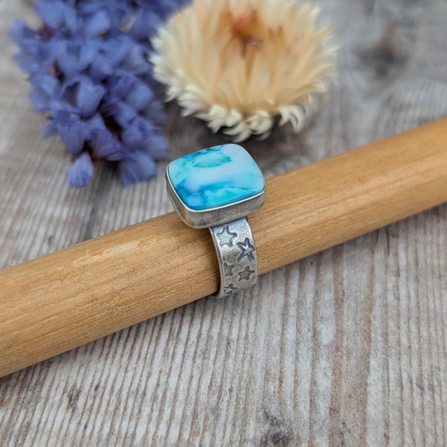 Blue Surfite Rectangle Ring with Silver Star Stamped Band resting on a wooden bar with purple and cream flowers in the background.