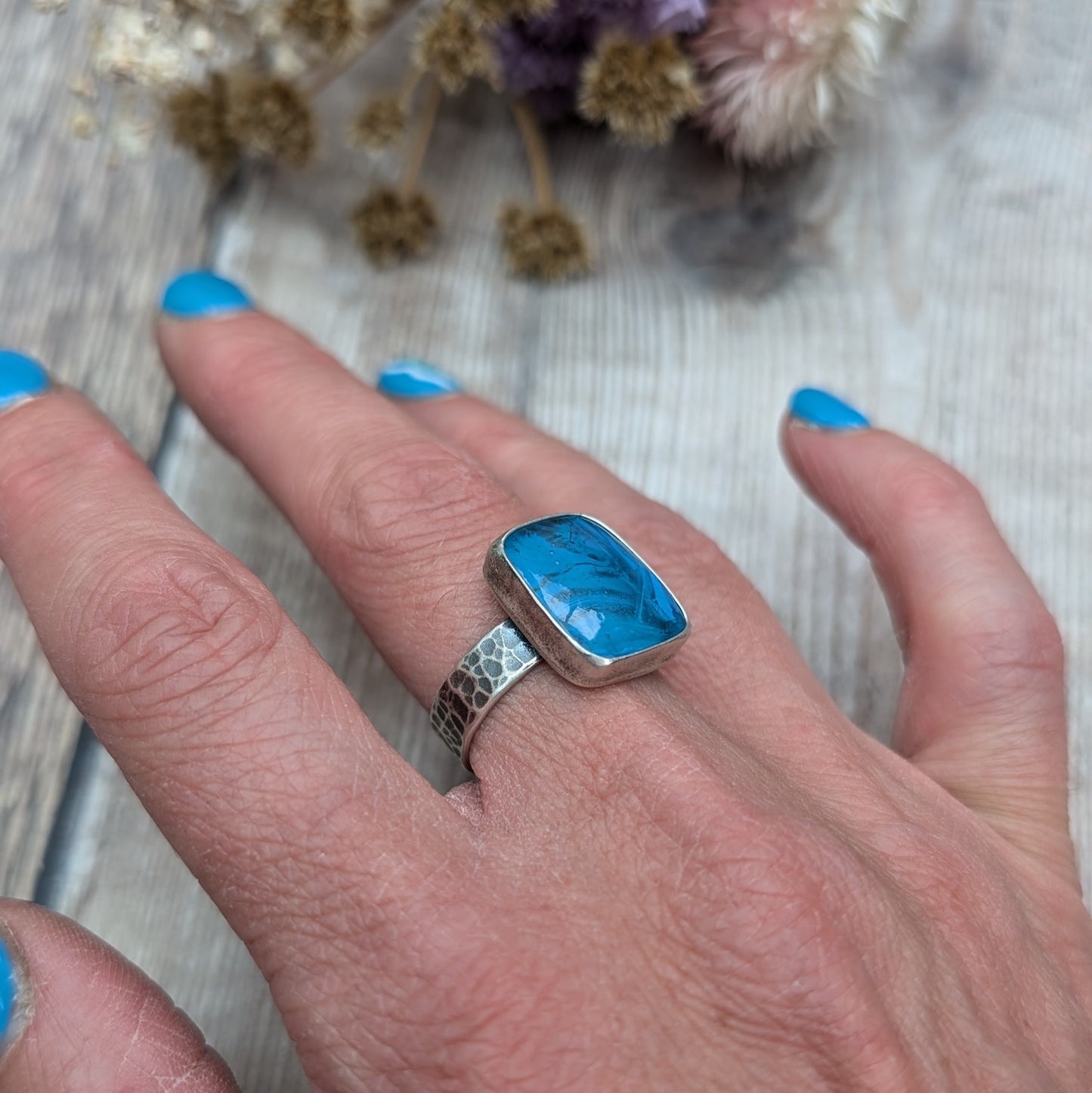 The ring features a rectangular-shaped blue gemstone with swirled patterns, set in a silver oxidised bezel and band with a textured design.