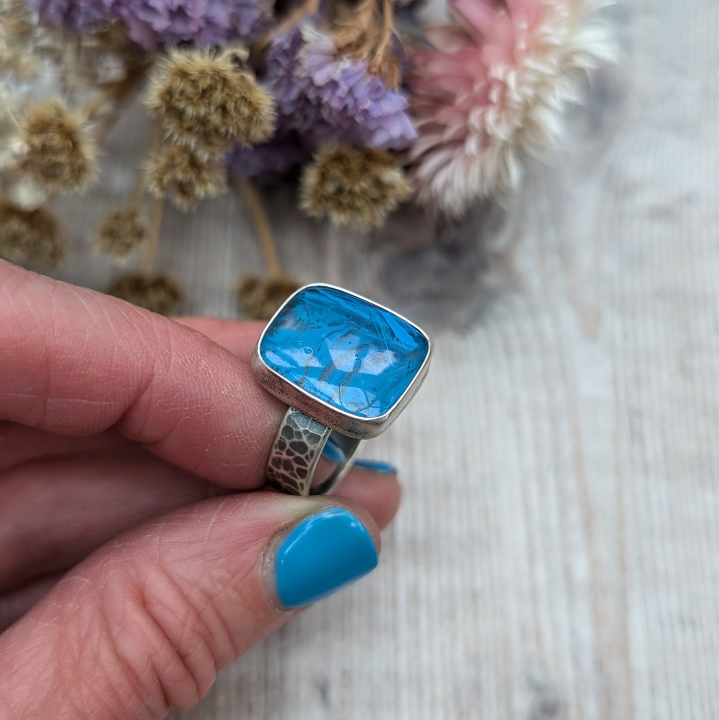 The ring features a rectangular-shaped blue gemstone with swirled patterns, set in a silver oxidised bezel and band with a textured design.