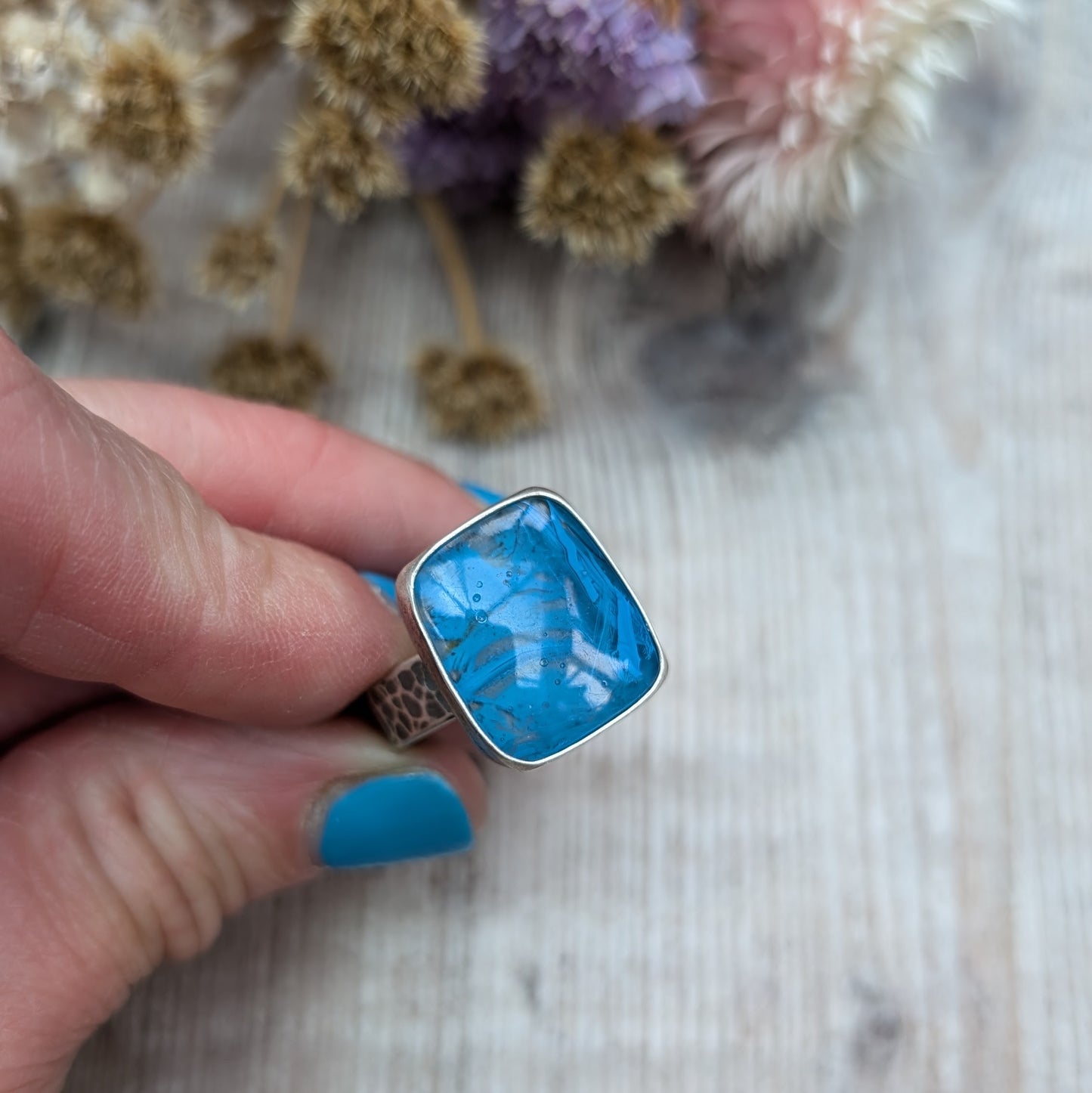 The ring features a rectangular-shaped blue gemstone with swirled patterns, set in a silver oxidised bezel and band with a textured design.