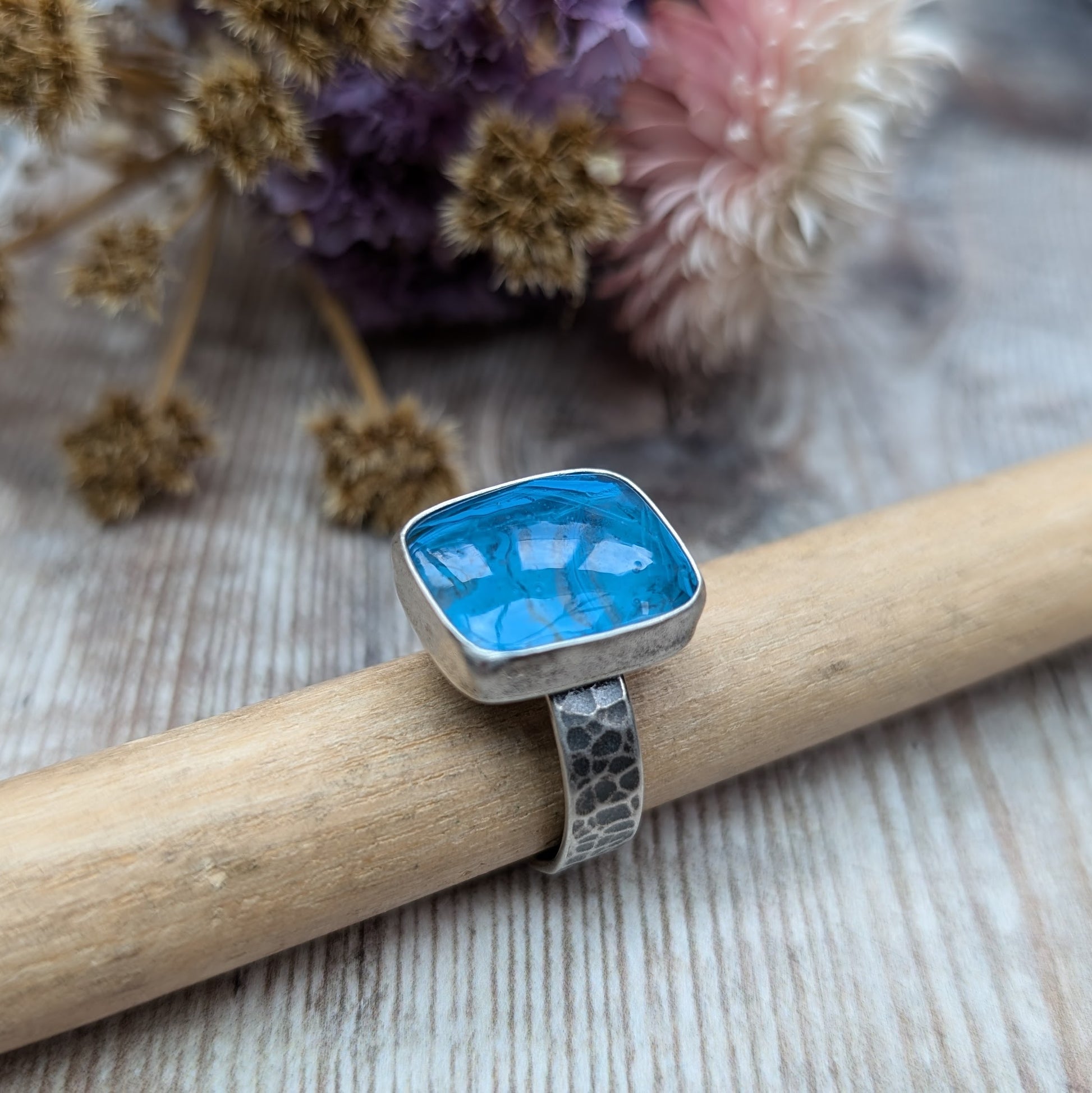 The ring features a rectangular-shaped blue gemstone with swirled patterns, set in a silver oxidised bezel and band with a textured design.