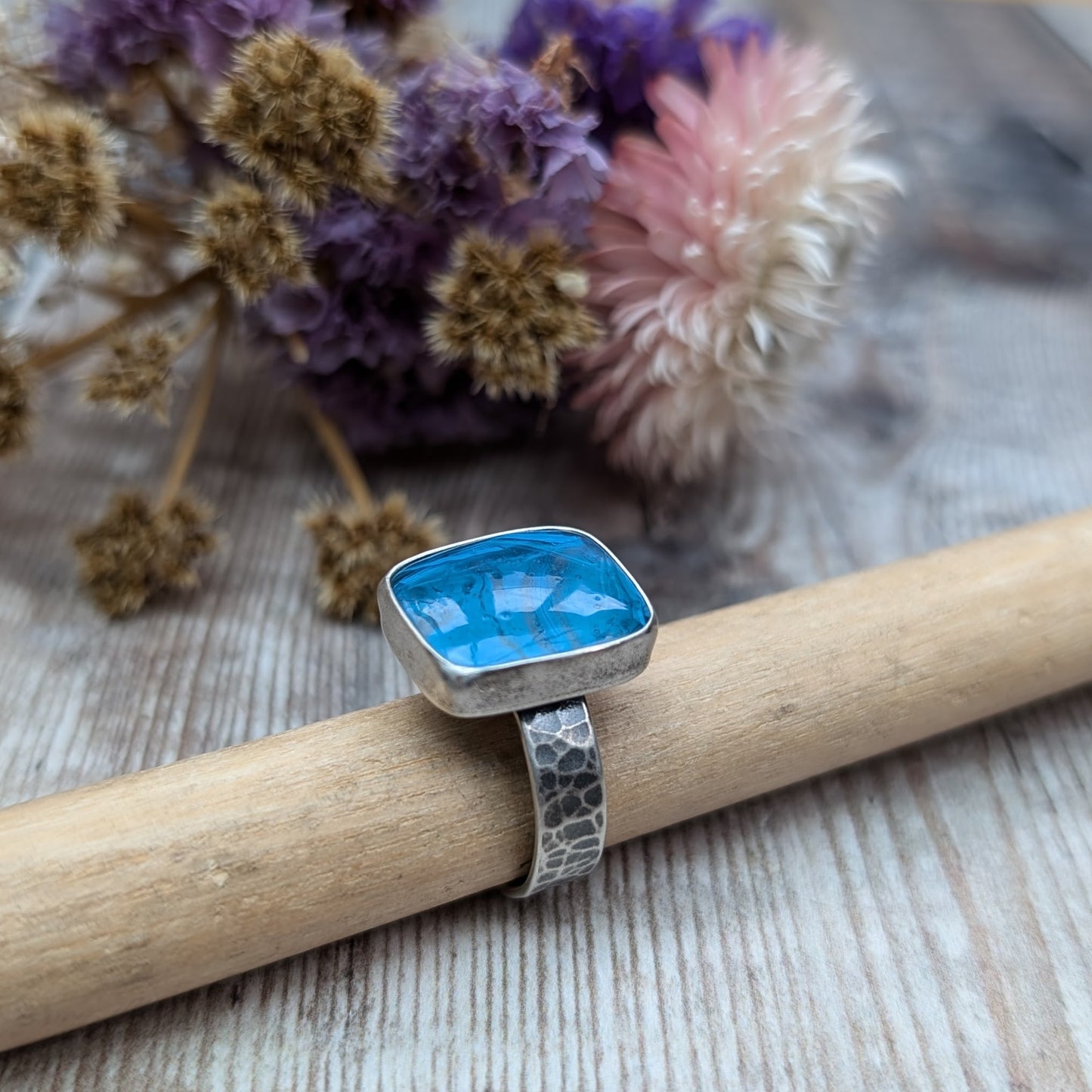 The ring features a rectangular-shaped blue gemstone with swirled patterns, set in a silver oxidised bezel and band with a textured design.