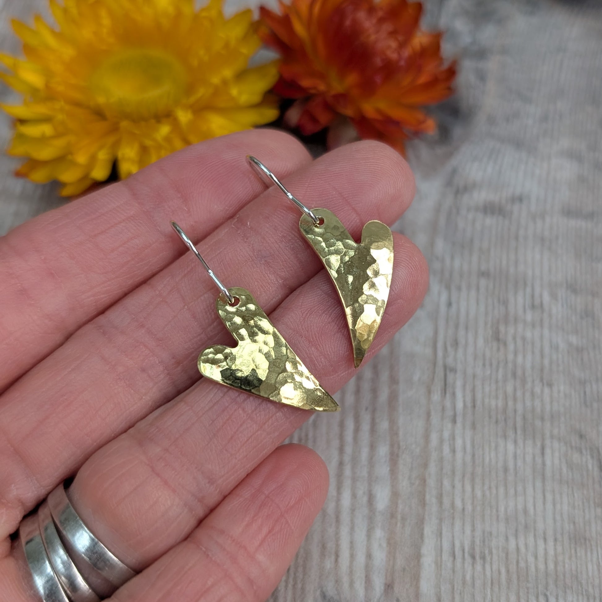 A hand holding the brass hammered curved heart earrings with orange and yellow dried flowers in the background, showing the hammered texture and curved heart shape.