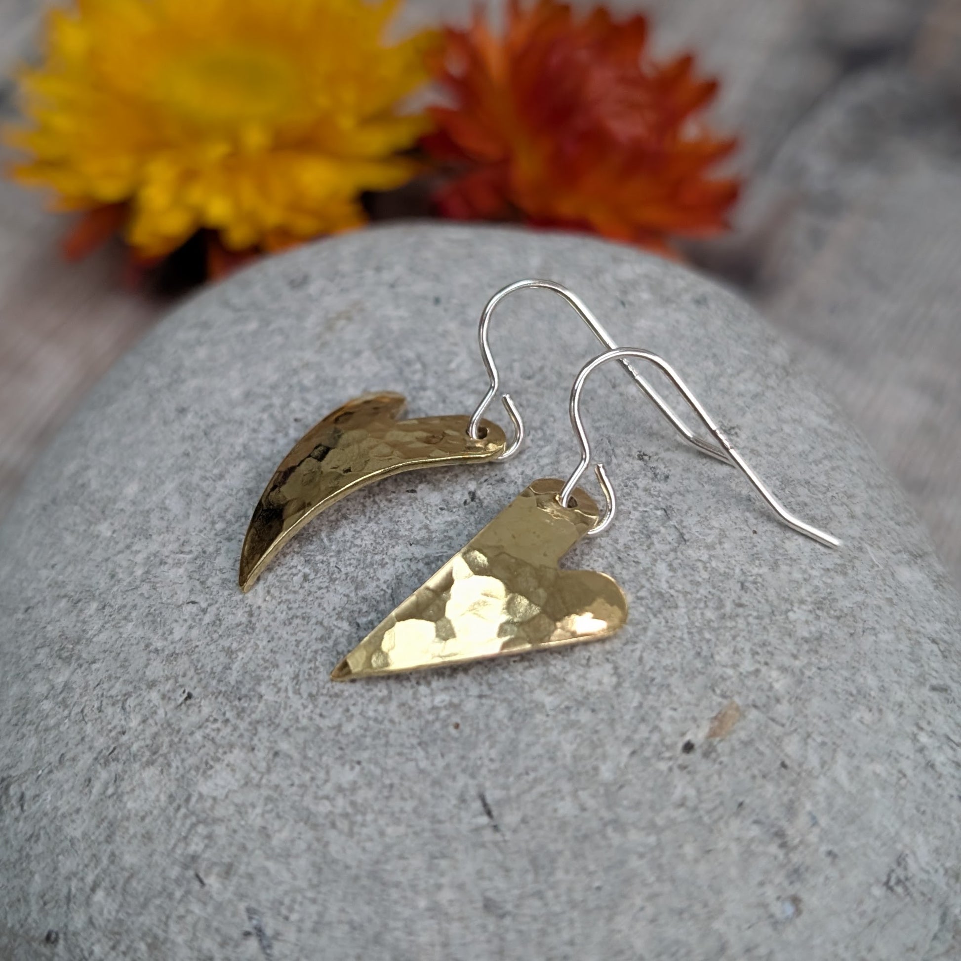 A pair of brass hammered curved heart earrings resting on a smooth grey stone with yellow and orange dried flowers in the background.