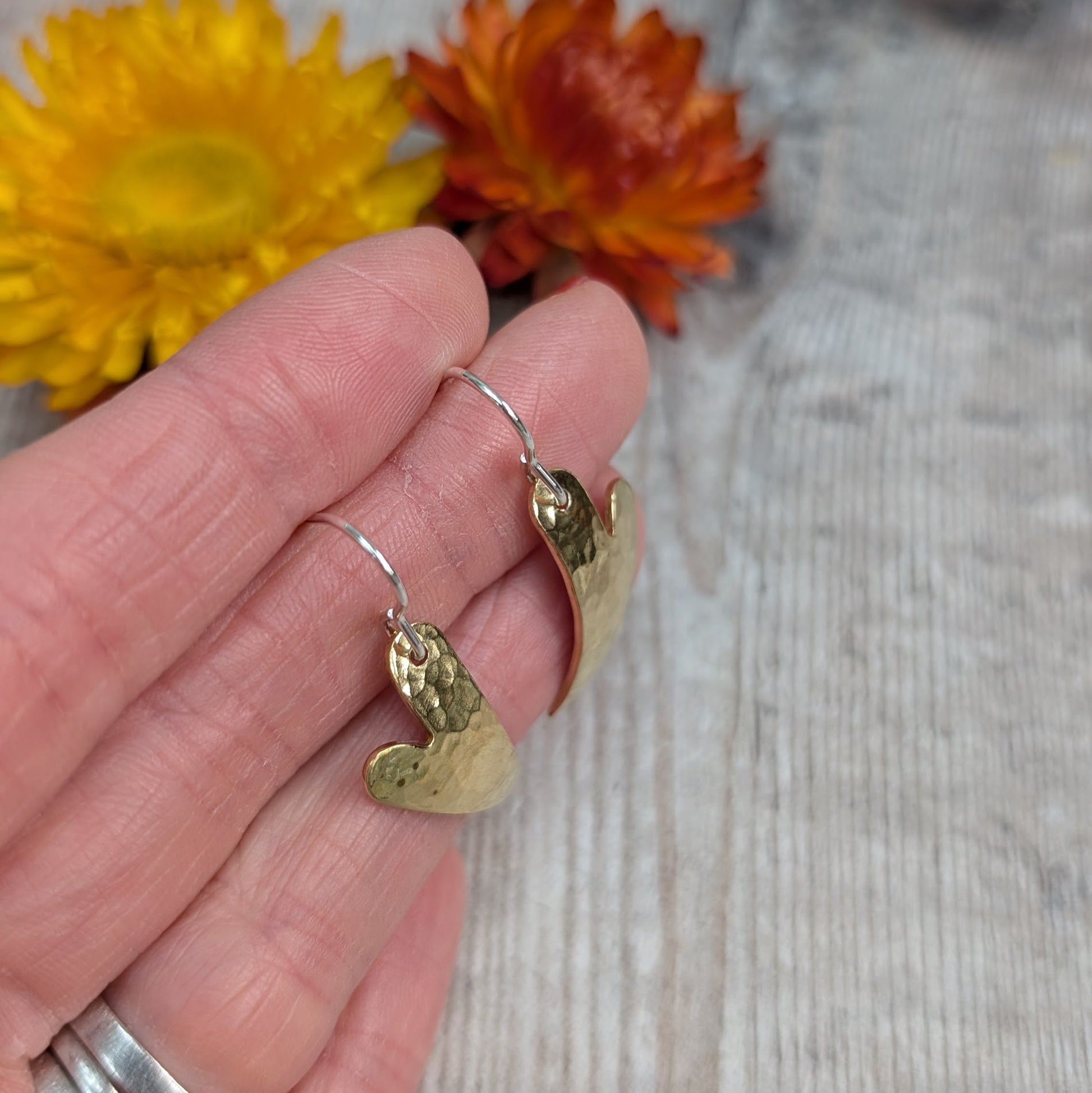 A hand holding the brass hammered curved heart earrings, showing their curved shape, hammered surface and silver ear hooks.