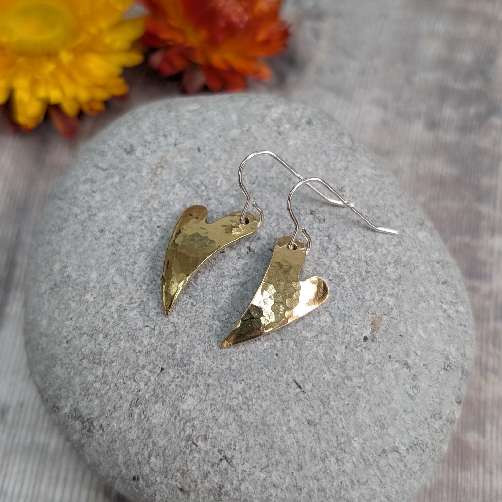 The brass hammered curved heart earrings displayed on a grey stone, highlighting their pointed heart shape, curved form and textured brass finish.