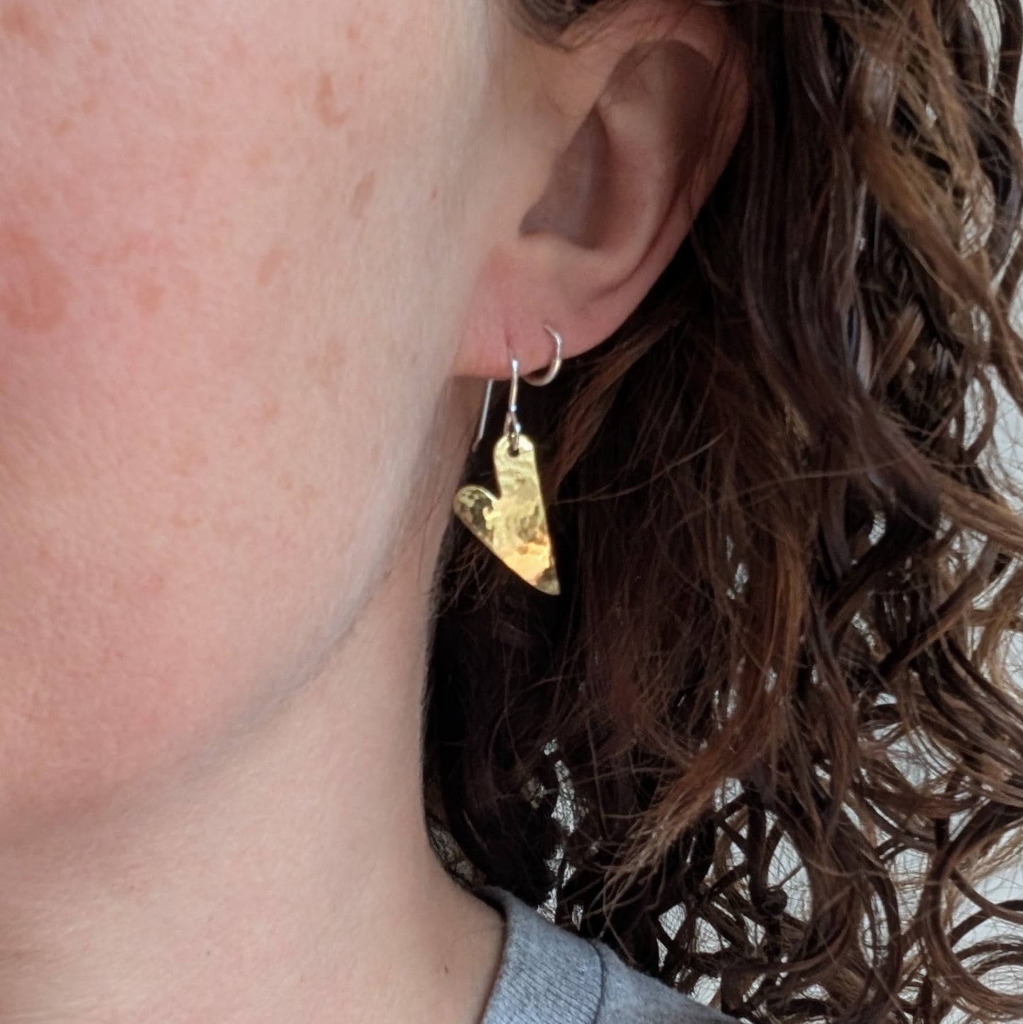 Close-up of one brass hammered curved heart earring being worn, showing the curved brass heart hanging from a silver ear hook.
