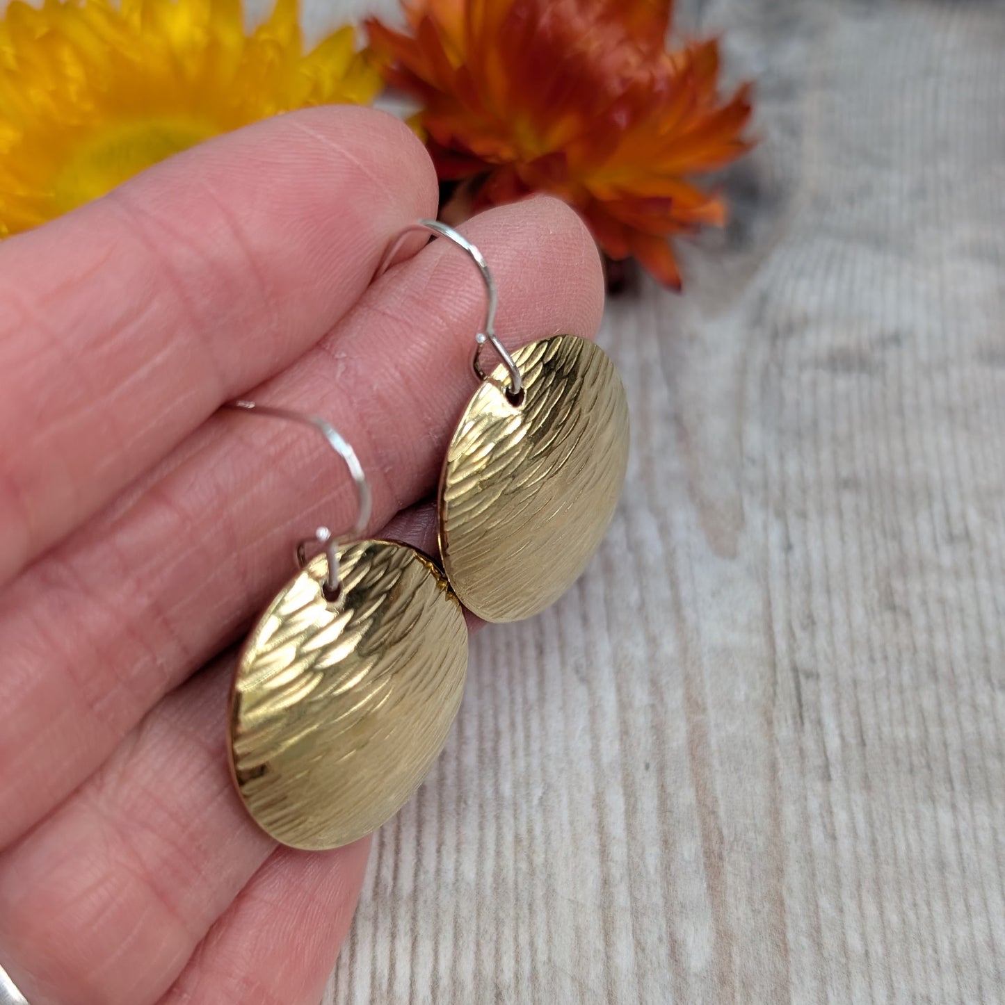 A close-up of a hand holding a pair of brass domed disc earrings with a hammered surface and sterling silver hooks, with yellow and orange flowers blurred in the background.
