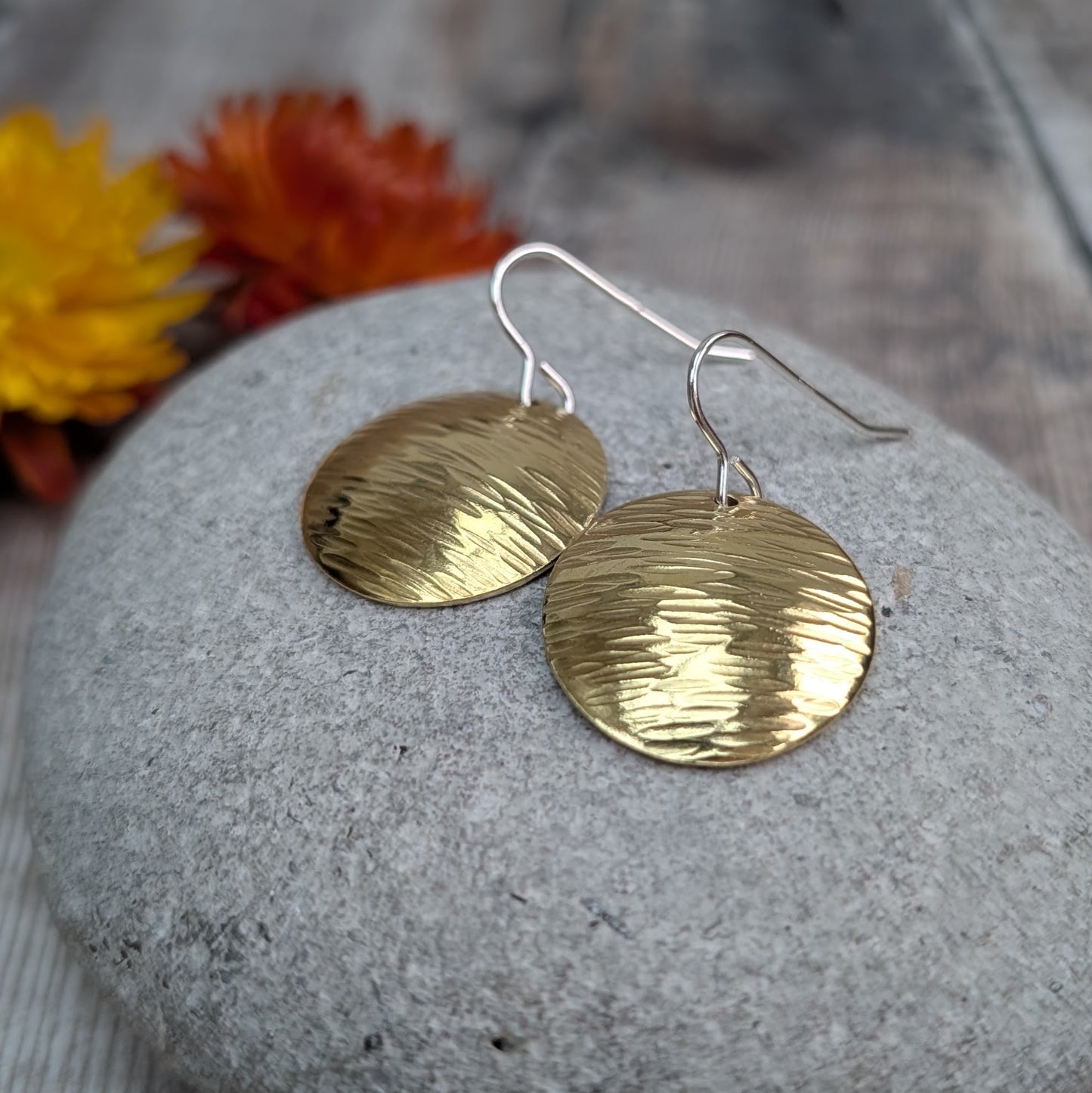 Two hammered brass domed disc earrings resting on a smooth grey pebble, with sterling silver hooks and autumn-coloured flowers in the background.